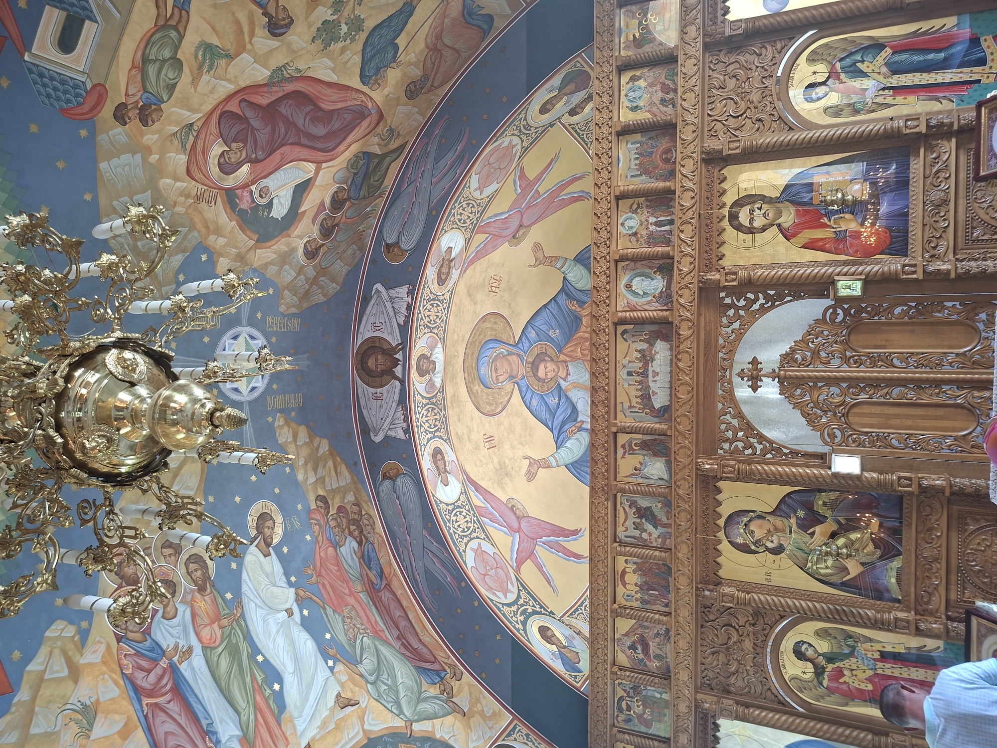 The image depicts the interior of a church, likely a Romanian Orthodox church, with extensive religious iconography. The ceiling is adorned with a mural depicting biblical scenes and figures, possibly angels and saints, rendered in a traditional style with vibrant colors and gold accents. A large, ornate brass chandelier hangs from the ceiling. The focal point of the lower section of the image is a wooden iconostasis, a wall of icons, separating the nave from the sanctuary. The iconostasis features intricately carved wooden details and numerous icons, including depictions of Jesus Christ, the Virgin Mary with the infant Jesus, and possibly saints or biblical events. Candles are visible in front of some of the icons, indicating active worship. A man wearing a light-colored shirt is partially visible in the foreground, looking downwards, suggesting he is participating in a service or observing the icons. Another person, partially visible from behind, wears a pink headscarf. The overall impression is one of a richly decorated and spiritually significant space. This specific imagery is consistent with the interior of the Saint Nicholas Church in Condrița, Moldova, known for its traditional Orthodox architecture and art.