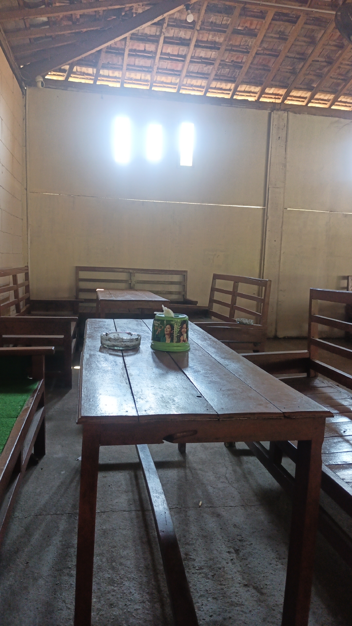 This is a medium shot, eye-level, interior photograph of a dimly lit, open-air seating area that appears to be a cafe or warung. The ceiling is constructed of exposed wooden beams and terracotta tiles. A bright light source, likely windows or skylights, is visible at the top of the back wall, casting a glare.
The main subject in the foreground is a weathered, dark wooden table with a glass ashtray and a green tissue box featuring images of women. The table is positioned on a concrete floor. Behind this table, further into the room, are more wooden tables and benches. The furniture is simple and rustic, made of unvarnished wood. Some of the benches have green cushions. The walls are painted a neutral, light color. The overall impression is one of a basic, functional outdoor or semi-outdoor seating area. The photograph was taken in Ngembalrejo, Indonesia.