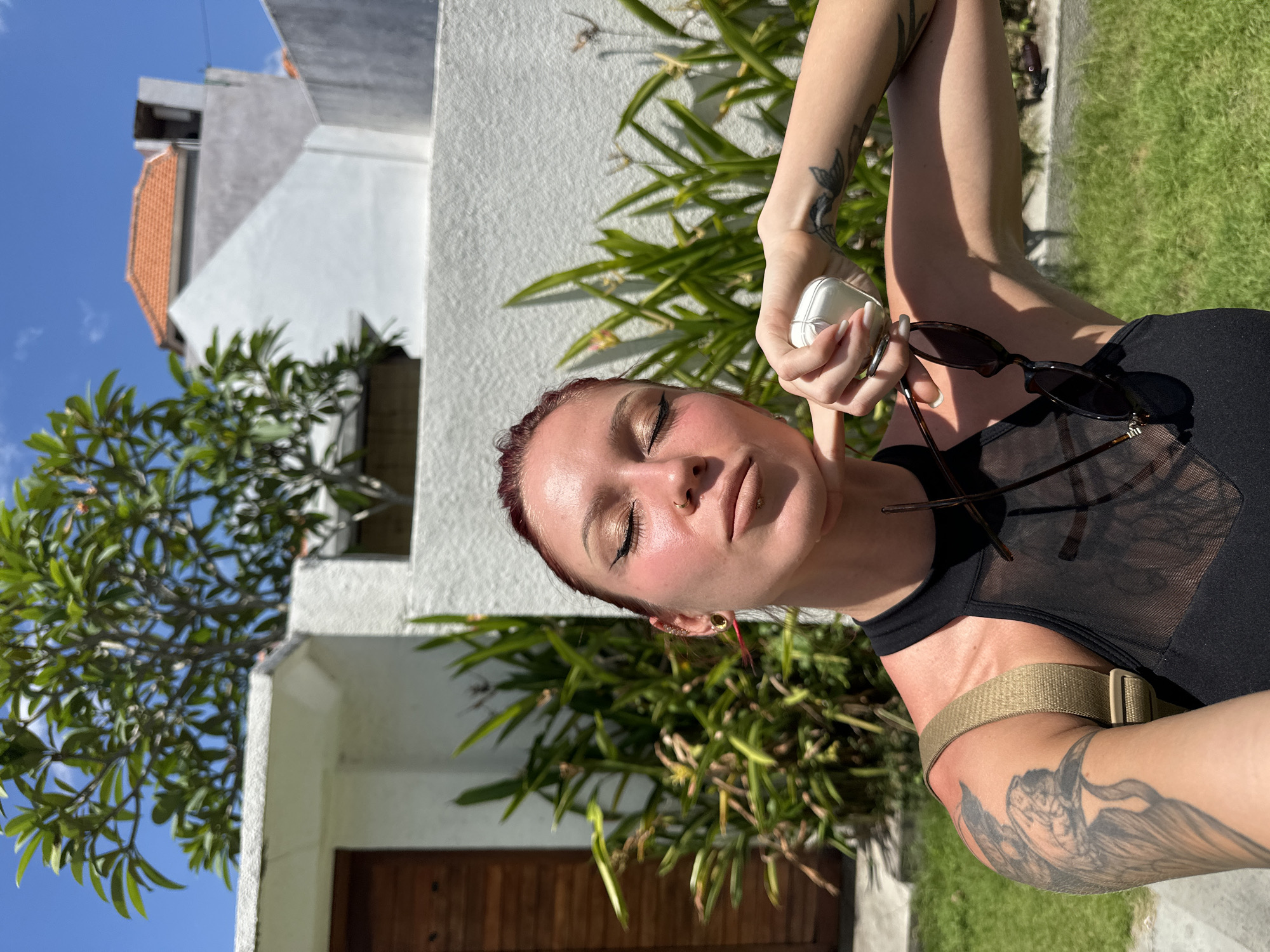 A light-skinned person with red hair is holding sunglasses and a white compact case in their right hand. The person is wearing a black top and a beige strap across their shoulder. They have tattoos on their arms. The background includes a white building with a wooden door, green plants, and a blue sky. The image was taken in Dalung, Indonesia.