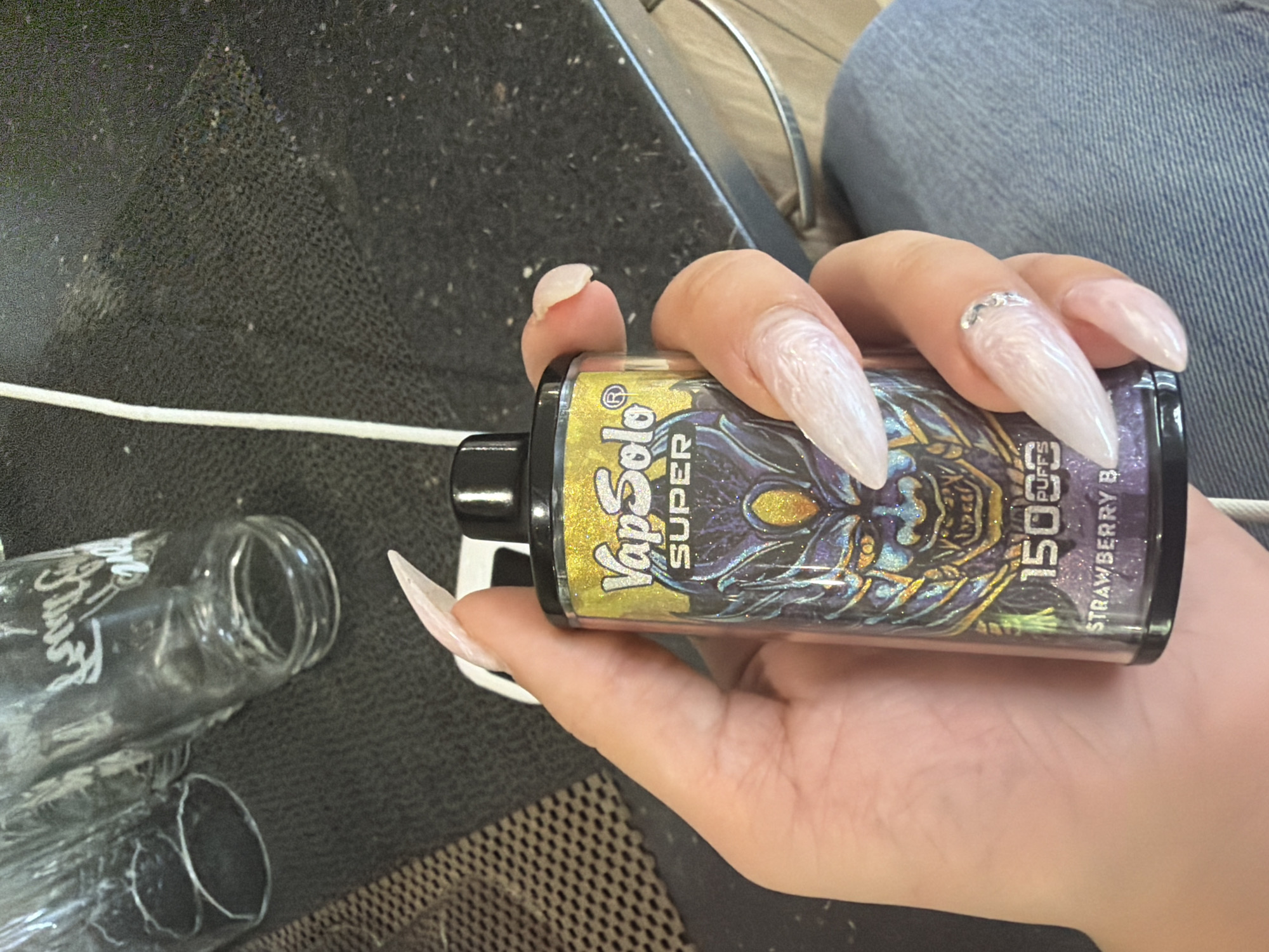 A close-up image shows a person's hand holding a vape device in an indoor setting. The hand has light skin and features long, pointed, light-colored acrylic nails, with one nail adorned with small, clear gems or glitter.
The vape device is black with a glittery, holographic design depicting a creature or skull. Visible text on the device includes "VapSolo SUPER," "15000 PUFFS," and "STRAWBERRY B" (likely indicating a strawberry flavor).
In the foreground, on a dark, textured surface, an empty clear glass bottle lies horizontally. A white cable runs across this surface, partially obscured by the bottle and the vape device. The background to the upper right shows a blurred section of what appears to be blue denim jeans, suggesting the person is seated.
The activity depicted is the holding of the vape device. Notable details include the prominent long acrylic nails and the specific branding and high puff count of the vape. The setting lacks specific visual cues for the stated location of Banya, Bulgaria. The time of day is not discernible from the artificial lighting.