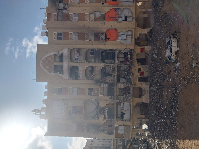 The image depicts a multi-story building in Khan Yunis, Palestinian Territory, exhibiting extensive damage. The structure, appearing to be residential or commercial, is light beige with a distinct arched architectural feature near the roofline. The entire façade is pockmarked with numerous small holes, consistent with shrapnel or bullet impacts.
Multiple floors show severe structural compromise: windows are shattered or entirely absent, revealing darkened and debris-filled interiors. Balconies on several levels are damaged, with railings distorted or missing. The second, third, and fourth floors from the bottom display the most significant destruction, with large sections of exterior walls collapsed, exposing internal rooms and twisted metal. On the right side of the building, orange tarpaulins cover openings on at least two different levels. Clothes are visible hanging on a damaged balcony on an upper-middle floor. The roof features water tanks and communication antennas.
At ground level, a vast expanse of pulverized concrete, rubble, and general debris surrounds the base of the building. Embedded within this debris field, towards the right foreground, lies the heavily damaged frame of a white vehicle, likely a car, with its roof caved in. The ground immediately in front of the building is bare earth, transitioning into the debris field. A strong sunlight source from the upper left casts a bright glare and creates a prominent shadow in the lower left corner of the frame. The sky is bright blue with scattered clouds. No individuals are visible in the scene.