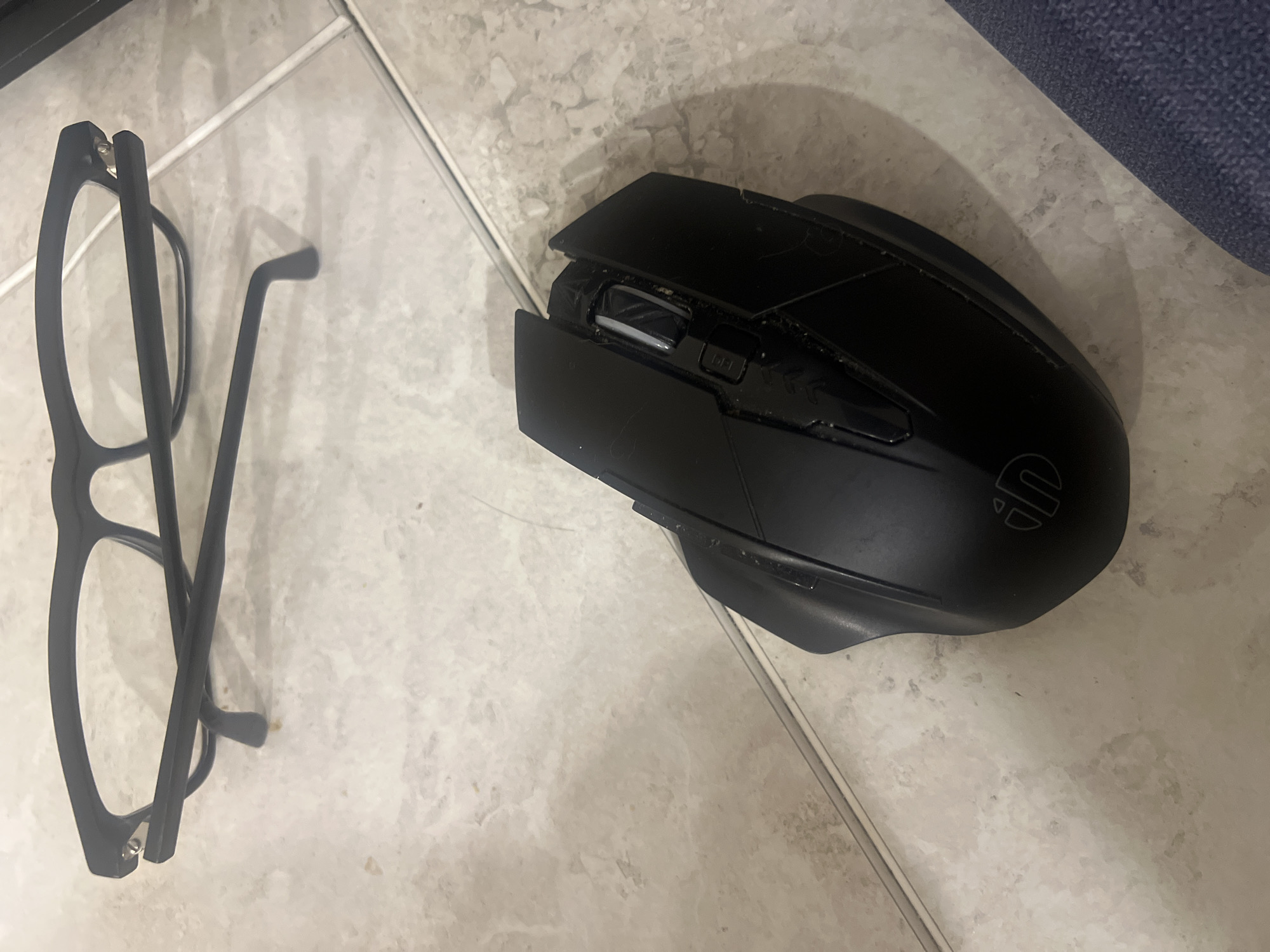A pair of black-rimmed glasses rests on a tiled floor. A black computer mouse is positioned in front of the glasses. The floor tiles are a light, marbled beige color. The lighting suggests an indoor environment. The scene is devoid of any human presence or specific landmarks that would indicate a location within Saratov, Russia.