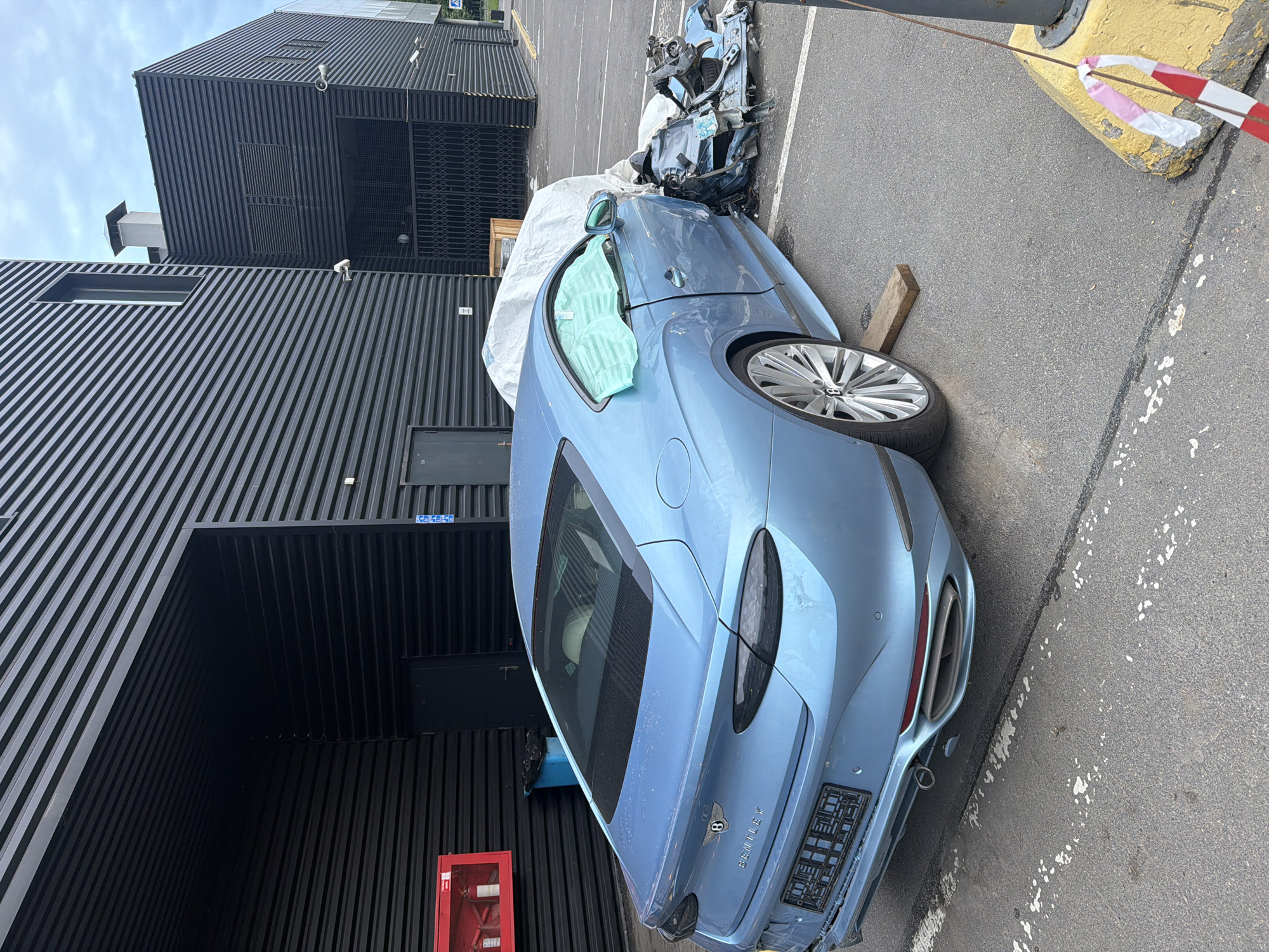 A heavily damaged blue Bentley Continental GT is parked in an asphalt lot outside a modern, black, corrugated metal building. The front passenger side of the car is severely impacted, with detached body panels and mechanical components scattered on the ground. The car is covered in a light-colored tarp, partially obscuring its rear. The building features large, dark entrances and security cameras are visible on its exterior. A red fire extinguisher box is mounted on the wall near one of the entrances. The ground is marked with parking lines and a yellow concrete bollard with red and white striped tape attached is present in the foreground. The sky is overcast.