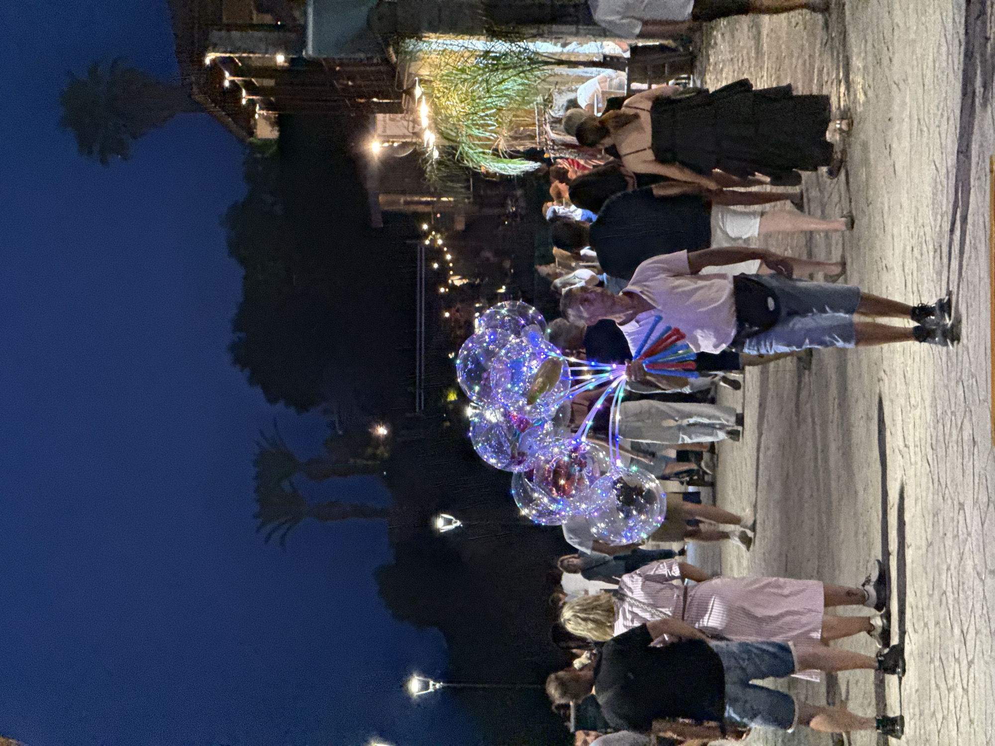 The image depicts a nighttime scene on a street in Kassandra Municipal Unit, Greece. The atmosphere is lively with a crowd of people walking along a cobblestone path. In the foreground, a man is holding a bunch of transparent balloons illuminated with colorful lights. He is wearing a light pink collared shirt, denim shorts, and black sneakers, with a black fanny pack around his waist. Other people in the vicinity are seen in casual attire, walking in various directions. The background reveals buildings with warm lighting, palm trees silhouetted against the dark sky, and streetlights casting a glow on the pathway. The overall impression is that of a bustling evening market or promenade.