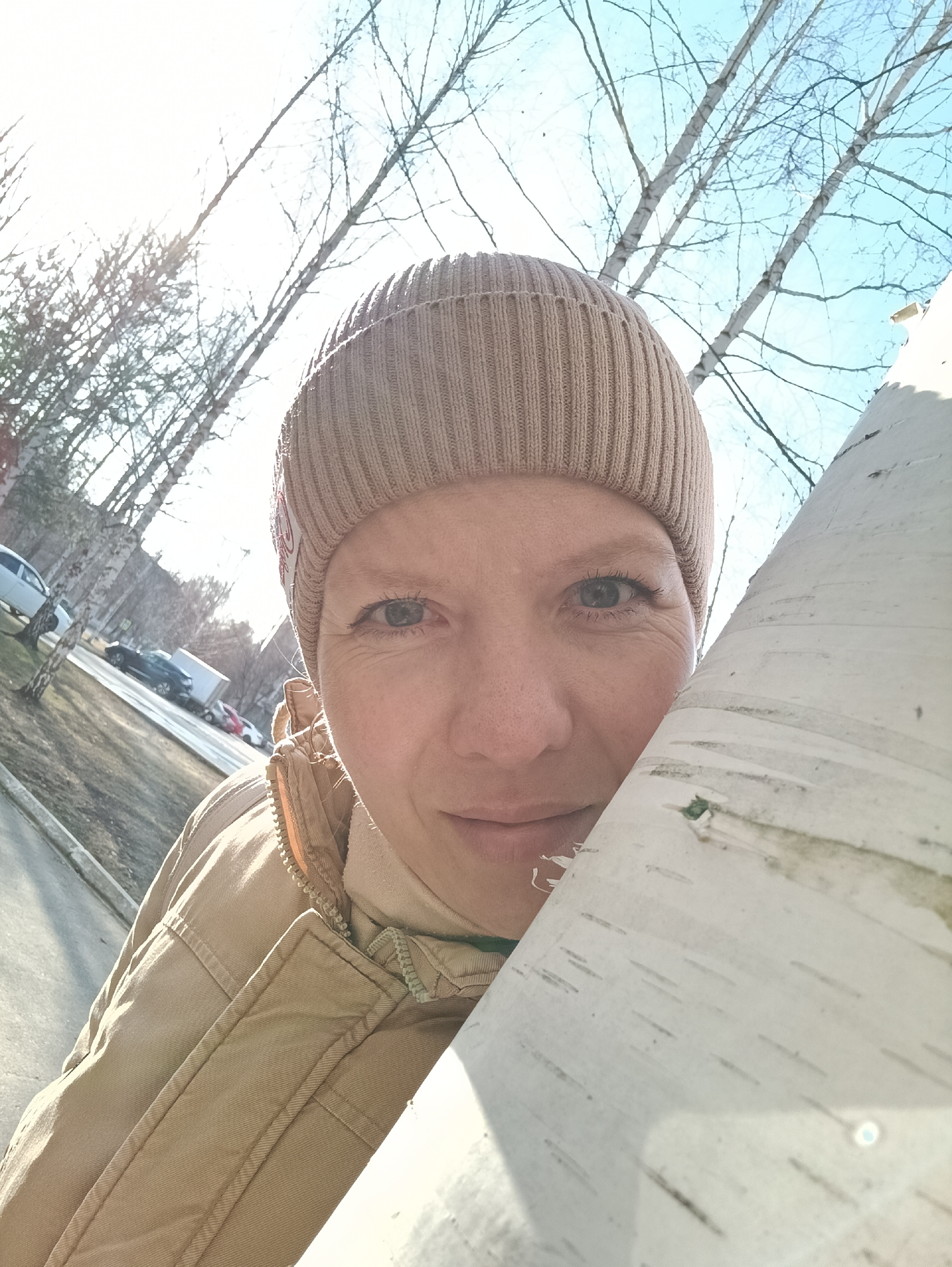 A close-up portrait captures a fair-skinned individual, wearing a tan ribbed beanie and a beige quilted jacket, partially obscured by the white bark of a birch tree. The person's face is centered, with clear blue eyes visible. Sunlight illuminates the left side of the frame, creating strong shadows and glare.
In the blurred background, a street scene unfolds. Several cars are parked along the sidewalk. Bare deciduous trees stand along the roadside. Some buildings are visible in the distance.
The scene is located in Chaikovsky, Russia, based on the presence of birch trees common to the region.