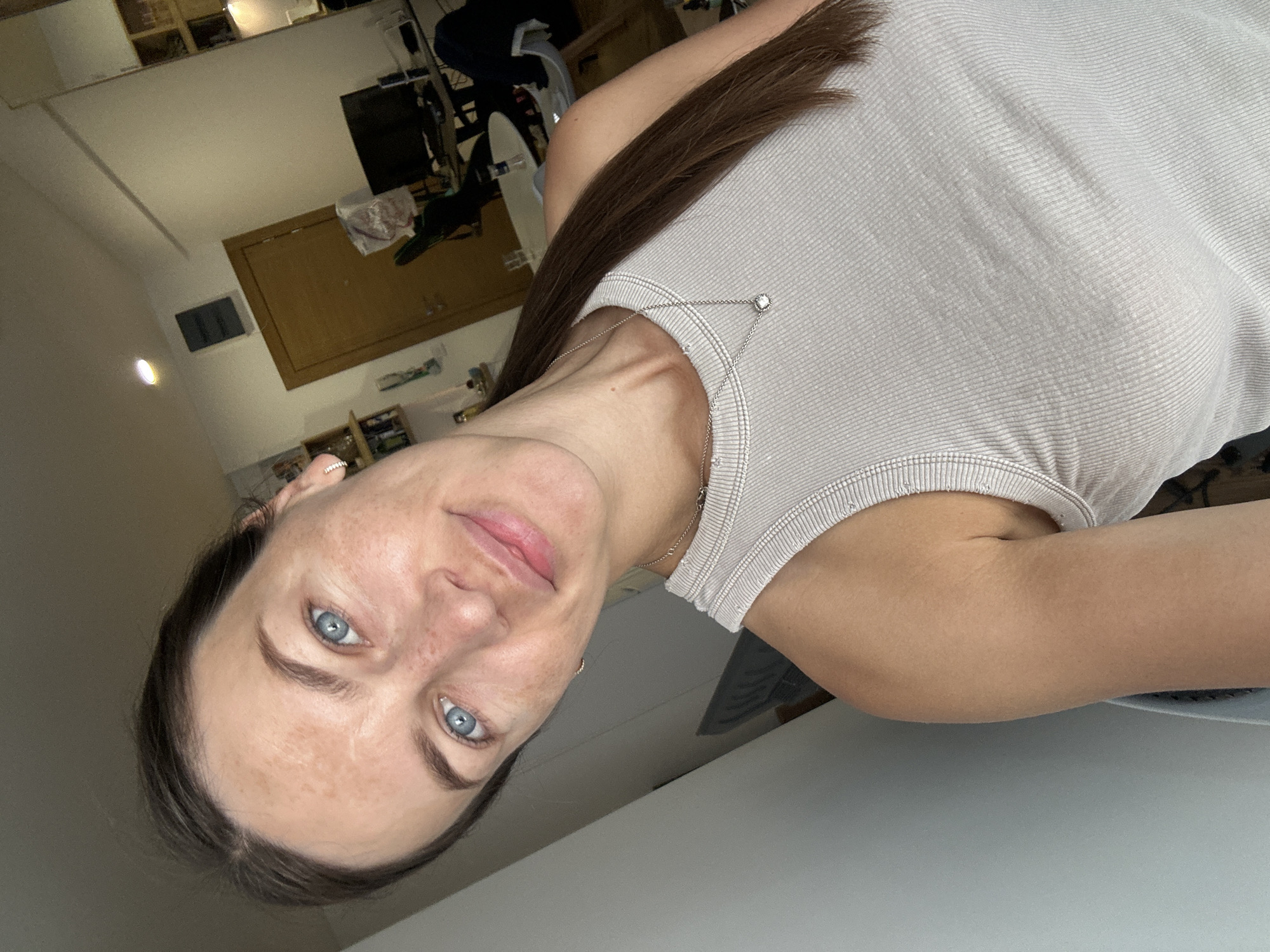 A woman with light skin and blue eyes is taking a selfie in a room. She has brown hair pulled back into a ponytail and is wearing a light-colored tank top with a silver necklace. The background appears to be an apartment with a door, a desk with a computer, and shelves filled with items. The lighting suggests it is daytime. The image is taken from a slightly upward angle, focusing on the woman's face and upper body.