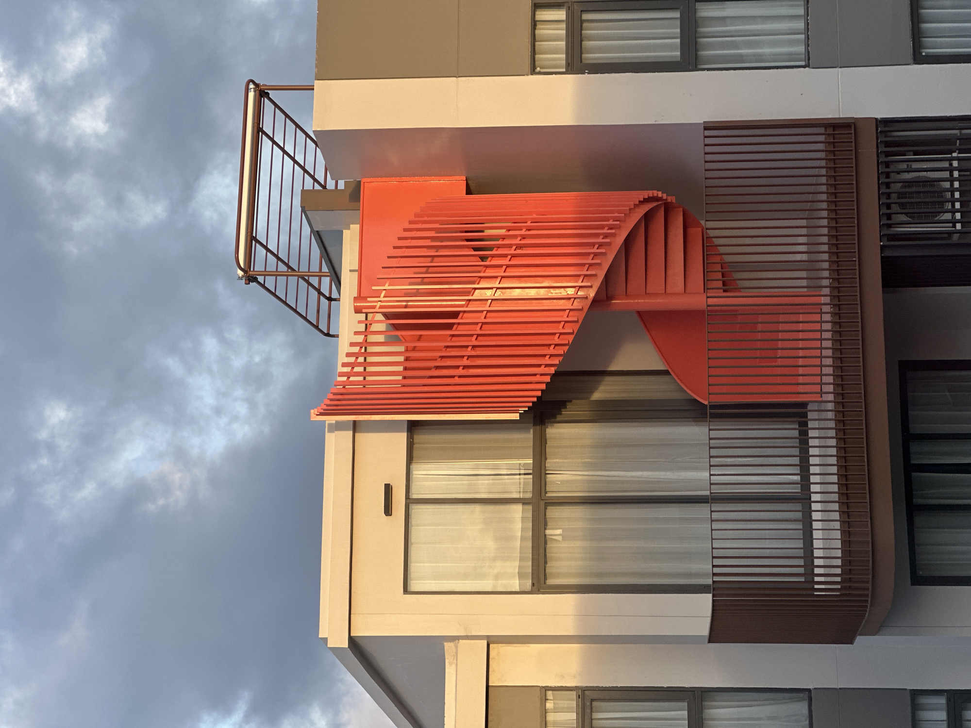 A modern building with a bright red spiral staircase against a cloudy sky is pictured in Choeng Thale, Thailand. The building features large windows with sheer white curtains, some reflecting the sky and sunlight. A brown metal railing encloses a balcony. The architecture suggests a contemporary residential or commercial property. The sky is overcast with dramatic, textured clouds.