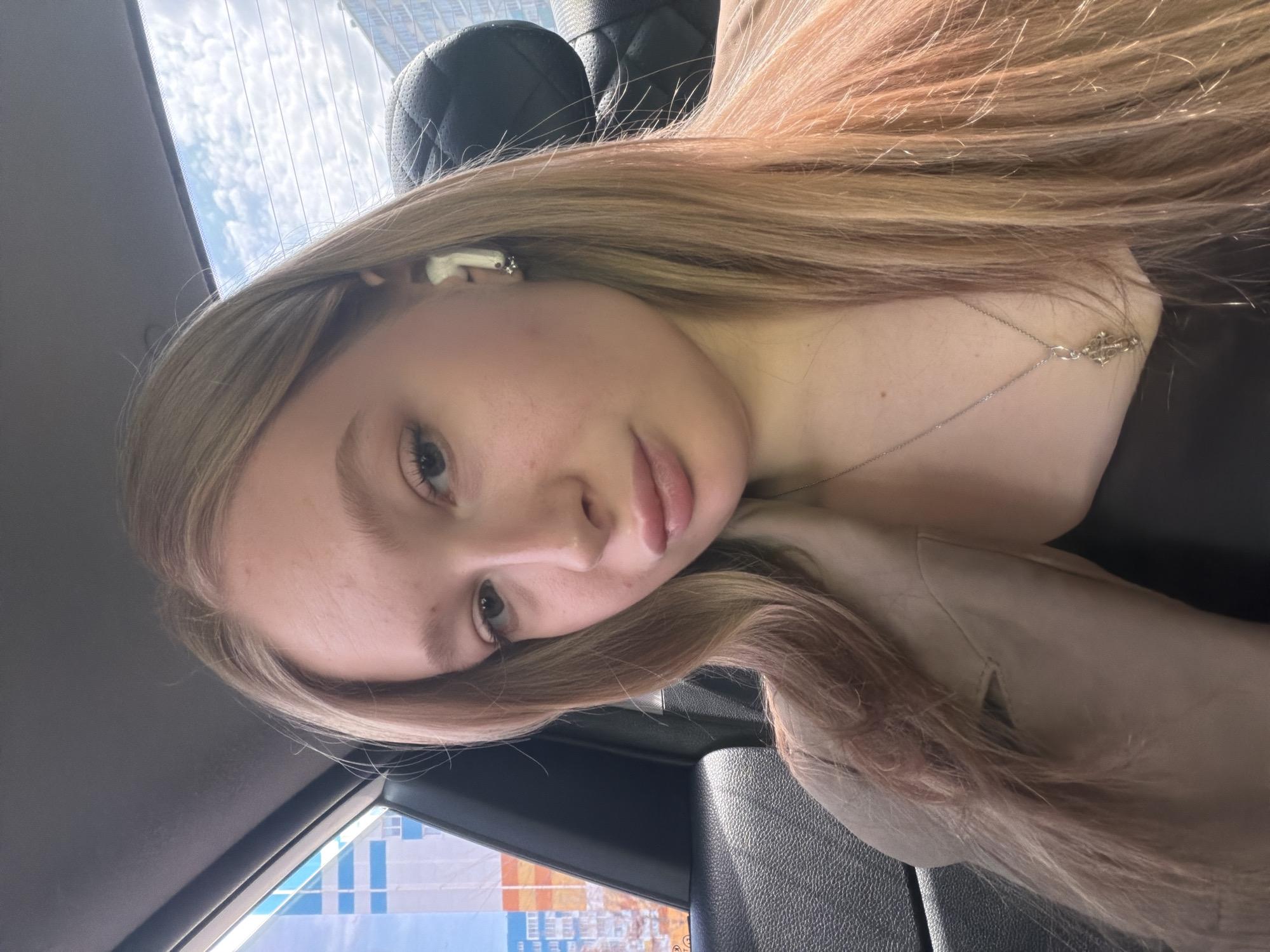 A fair-skinned young woman with blonde hair is visible from the shoulders up, likely in the backseat of a car. She is facing the camera with her head tilted to the left. She is wearing what seems to be a light brown jacket and a silver necklace. There is a white earpiece in her left ear. Outside the car window, there are buildings, suggesting an urban environment. Given that the location is Засечное, Russia, it is possible that the buildings seen out of the window are in or near that city. The image appears to be a selfie taken inside a vehicle. The scene seems unremarkable, capturing a casual moment.