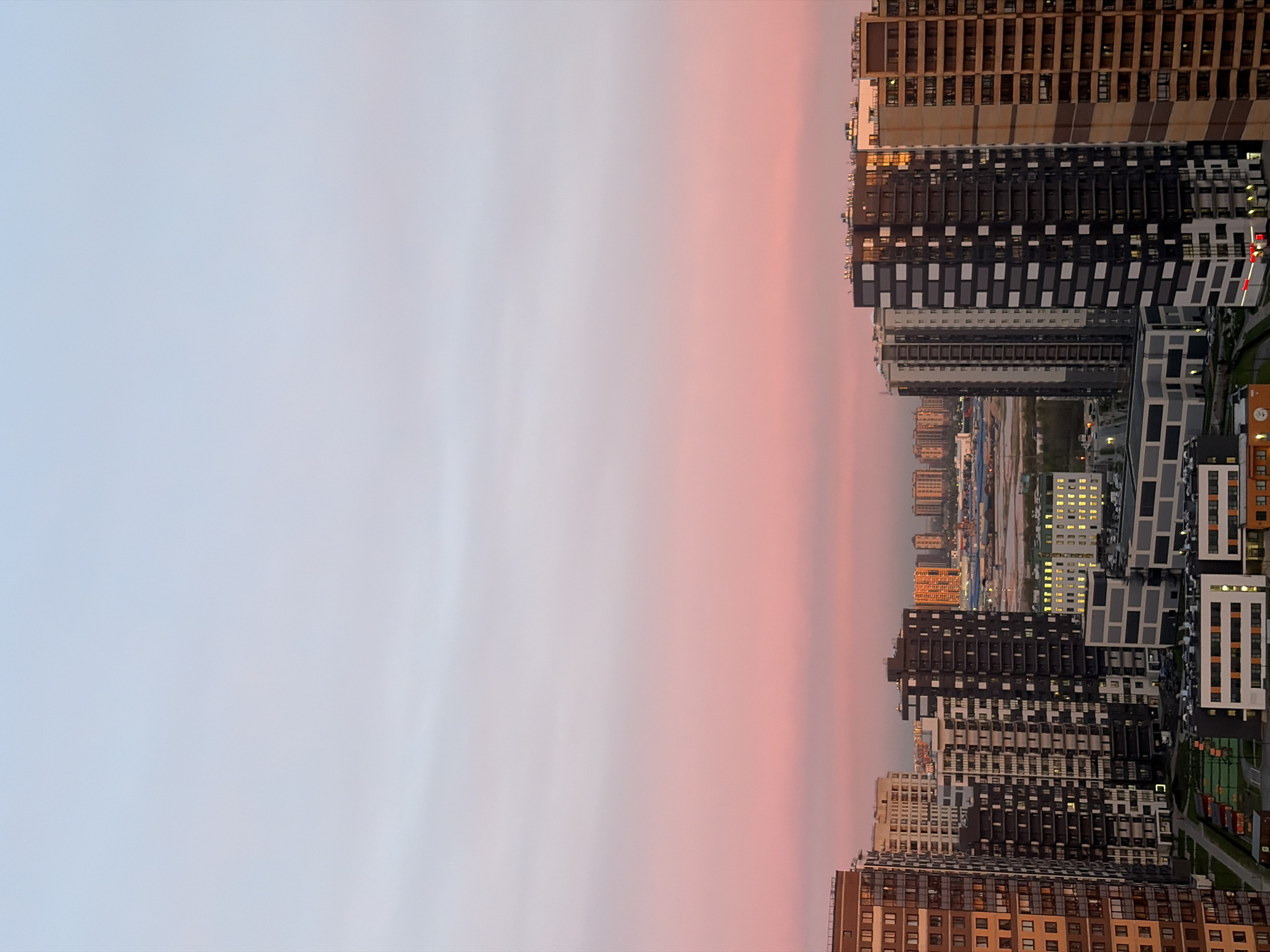 The image is an aerial view of a cityscape at dusk or dawn, likely in Saint Petersburg, Russia. High-rise buildings dominate the right side of the frame, varying in color from brown and beige to white and dark gray. Many of the buildings have lit windows. The skyline recedes into the distance. The buildings align against a gradient of light blue to pink sky. No people are visible. The scene is serene.
