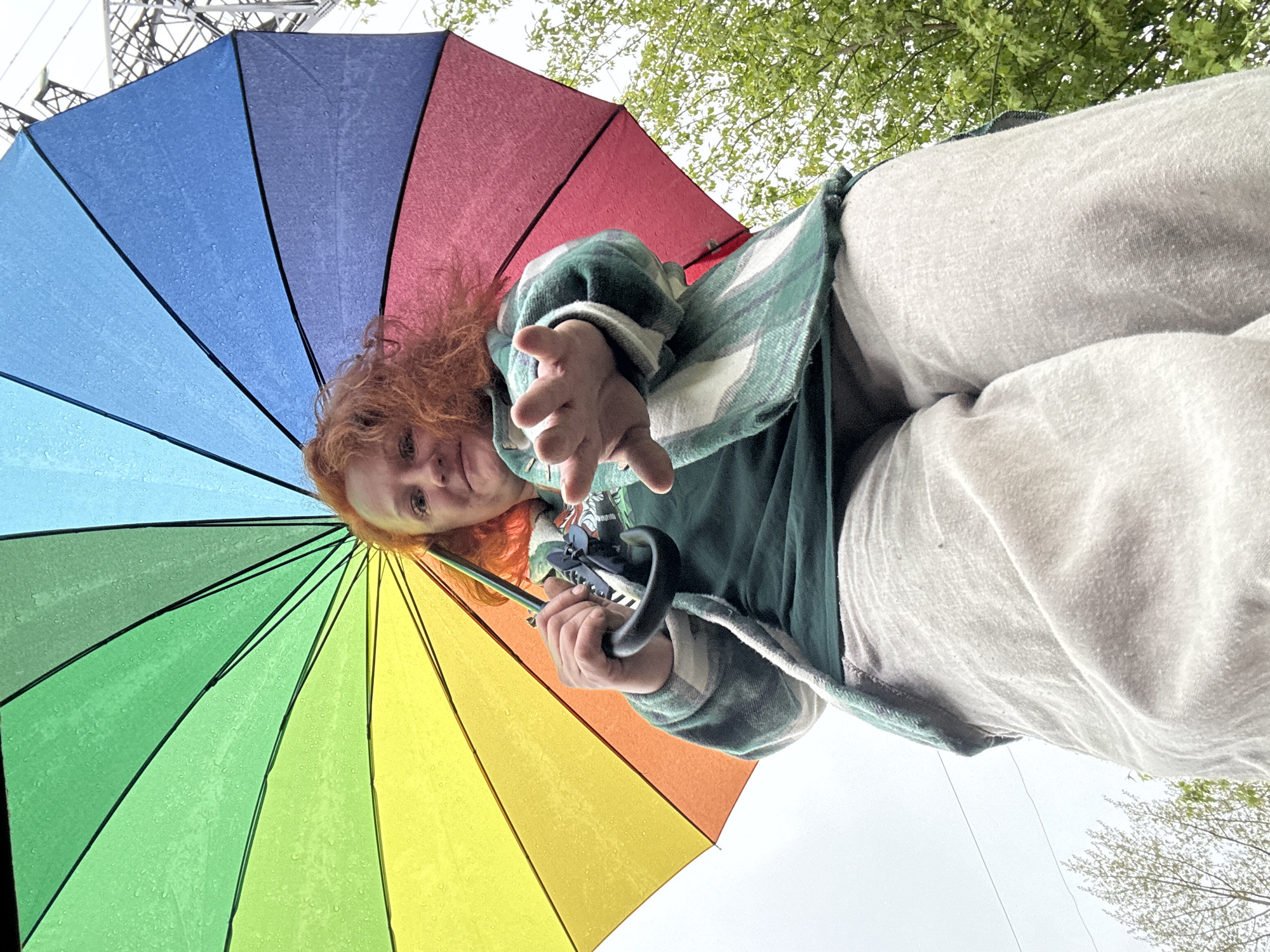 A low-angle shot shows a person with red hair holding a multi-colored umbrella. The umbrella is divided into rainbow sections and is wet. The person is wearing a green plaid shirt and light gray pants. They are holding out their hand towards the viewer. Overhead, there is a power grid. The background includes a sky obscured by clouds and tree branches, suggesting an outdoor environment. The location is Chaikovsky, Russia, based on additional information not present in the image.