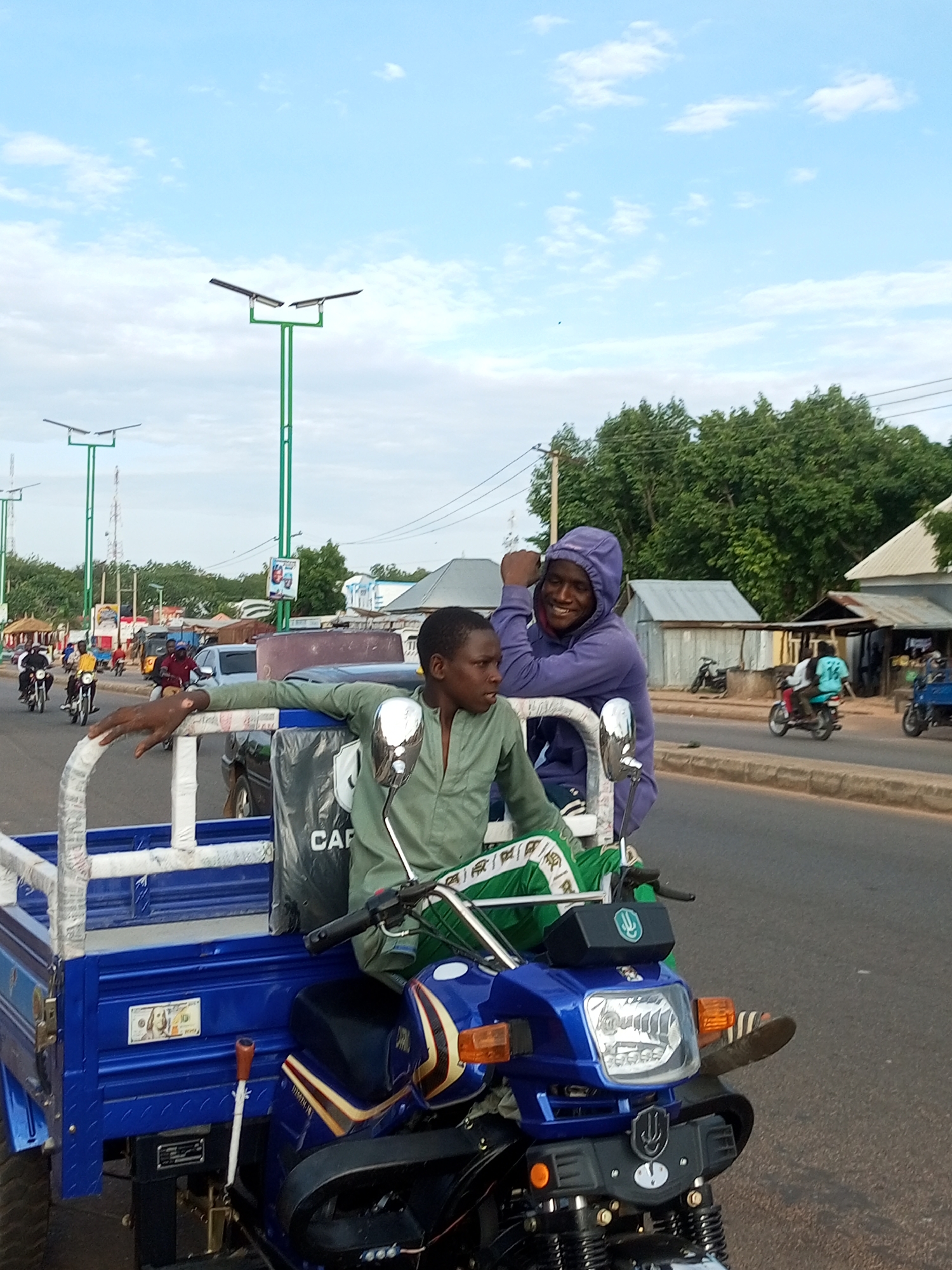 The image depicts an outdoor, daytime scene on a paved road in an unknown city. In the foreground, two males are positioned on a blue motorized tricycle, which features a cargo bed. The male on the left, in what appears to be the driver's position, wears a light green long-sleeved shirt and green and white patterned trousers. His left hand rests on the front railing of the tricycle. The male on the right, in a passenger position, wears a purple hooded sweatshirt with the hood up; he is smiling broadly and looking towards the camera, with his left hand on the railing and his right arm bent.
The tricycle's front section is blue, resembling a motorcycle, with a prominent headlight, orange turn signals, and a black seat. Two "U" shaped logos are visible: one on the handlebars and another below the headlight. A small wooden lever extends from the lower part of the vehicle. The blue metal cargo bed has a white railing, partially covered with white material. A rectangular sticker depicting a U.S. 100-dollar bill is affixed to the side of the cargo bed. A black plastic bag rests on the seat or cargo area behind the driver.
In the mid-ground and background, multiple vehicles, primarily motorcycles, are visible on the road, some with passengers. The road itself appears dry. Several tall, green streetlights topped with solar panels line the road on both sides. Overhead utility lines are also present. The background features various single-story buildings, some with corrugated metal roofs, alongside numerous green trees and other vegetation. The sky is bright blue with scattered white clouds.
