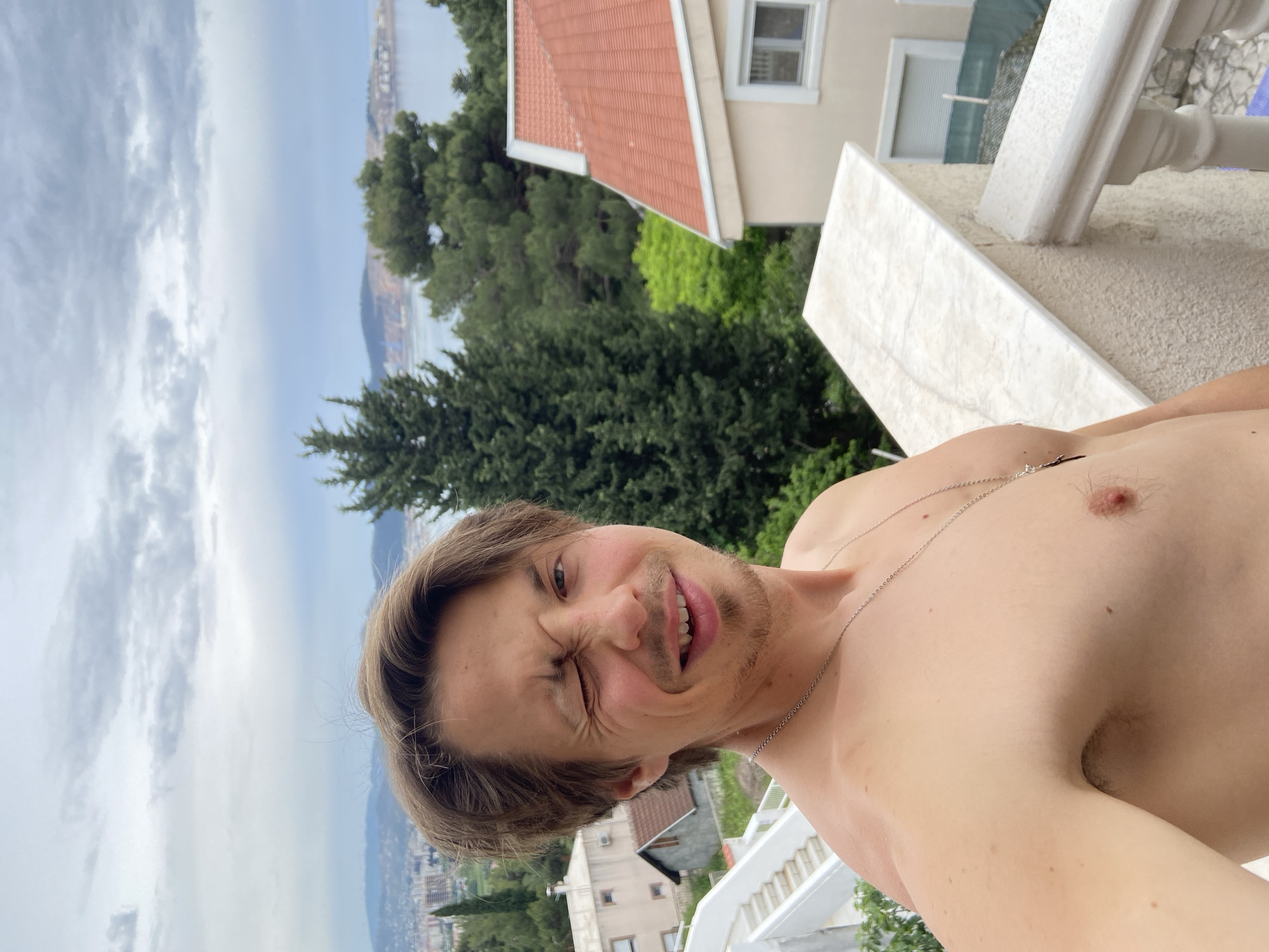 A shirtless man with light brown hair is standing on a balcony, making a comical face while looking directly at the camera. He is wearing a silver chain necklace. Behind him, there is a view of Šušanj, Montenegro, with residential buildings and lush green trees. The sky is cloudy. A building with a red tiled roof is visible to the upper right. Part of a white marble balcony railing is visible. The photo appears to be a selfie taken by the man.