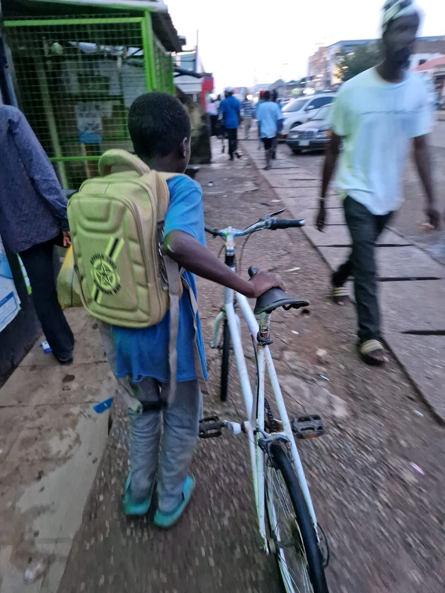 A young boy wearing a blue shirt and grey pants, carrying a khaki backpack, stands beside a white bicycle on a sidewalk in Jos, Nigeria. The boy is facing away from the camera, looking towards the right. The bicycle has a black seat and is positioned in the foreground, partially obscuring the boy's legs. Behind the boy, other pedestrians and vehicles are visible in the background, suggesting a street scene. The environment appears to be an urban setting with buildings and a road. The overall lighting indicates it might be late afternoon or early evening.