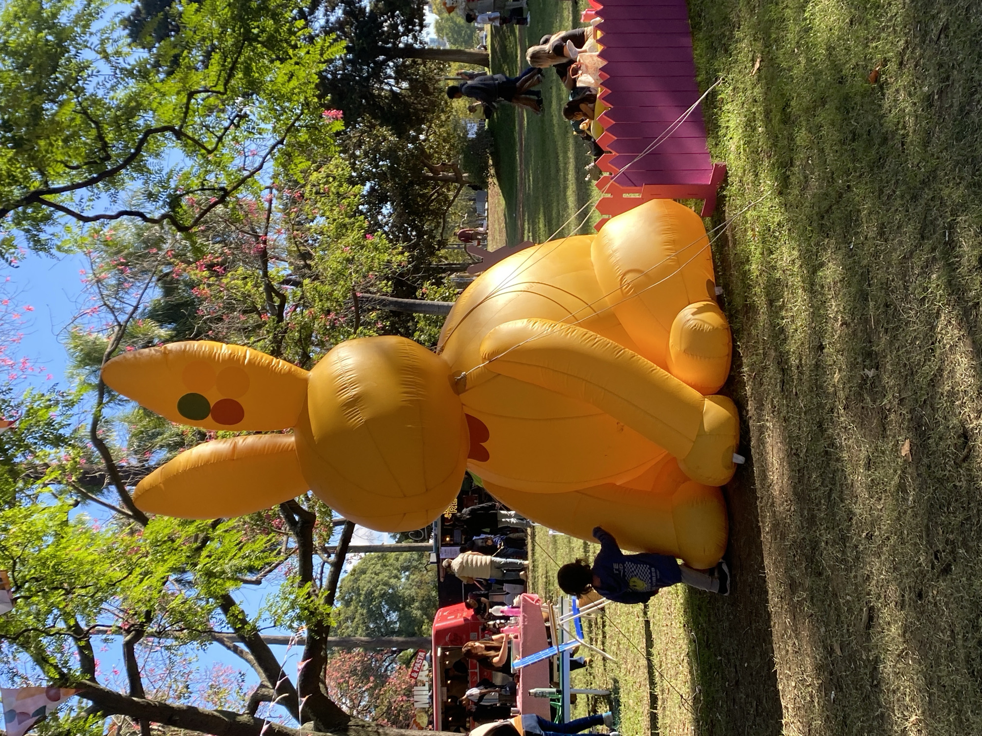 The image depicts an outdoor scene featuring a large, inflatable yellow bunny. The bunny is resting on a grassy area, tethered by ropes to a pink and purple fence. A small child in a blue jacket stands near the bunny's feet. In the background, there are trees, people, and a red food truck. The setting appears to be a park or public space. The location is the city of Buenos Aires, Argentina.