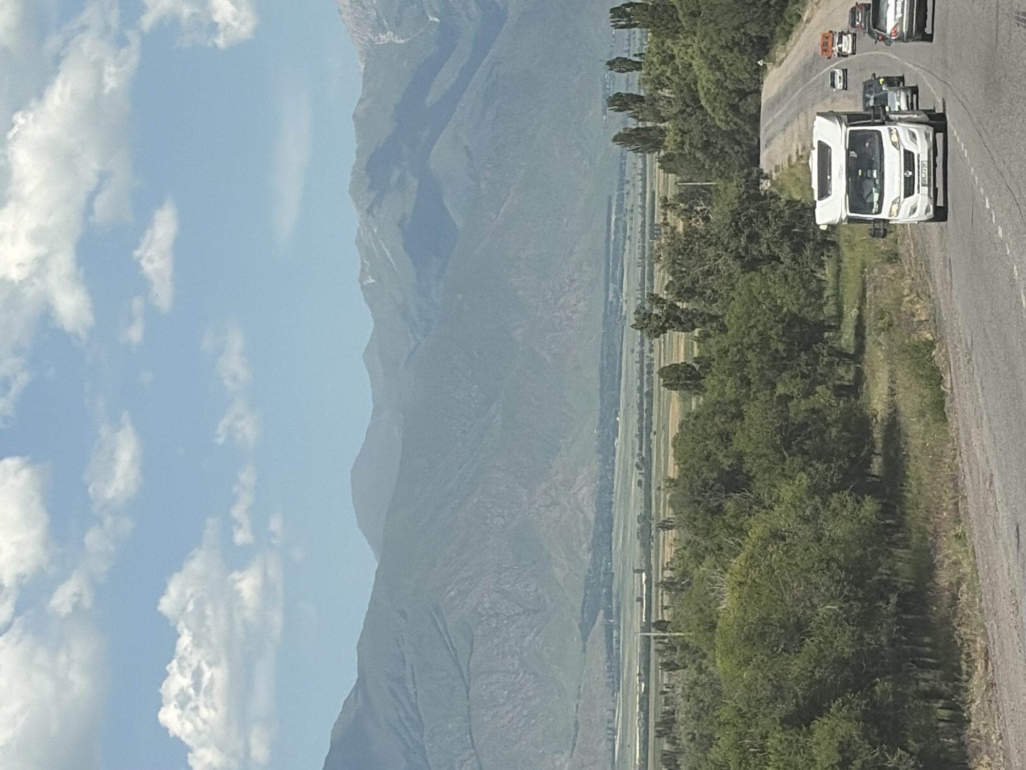 A white camper van is driving on a road with other cars behind it. The road is surrounded by trees and in the background there are mountains. The sky is blue with white clouds.