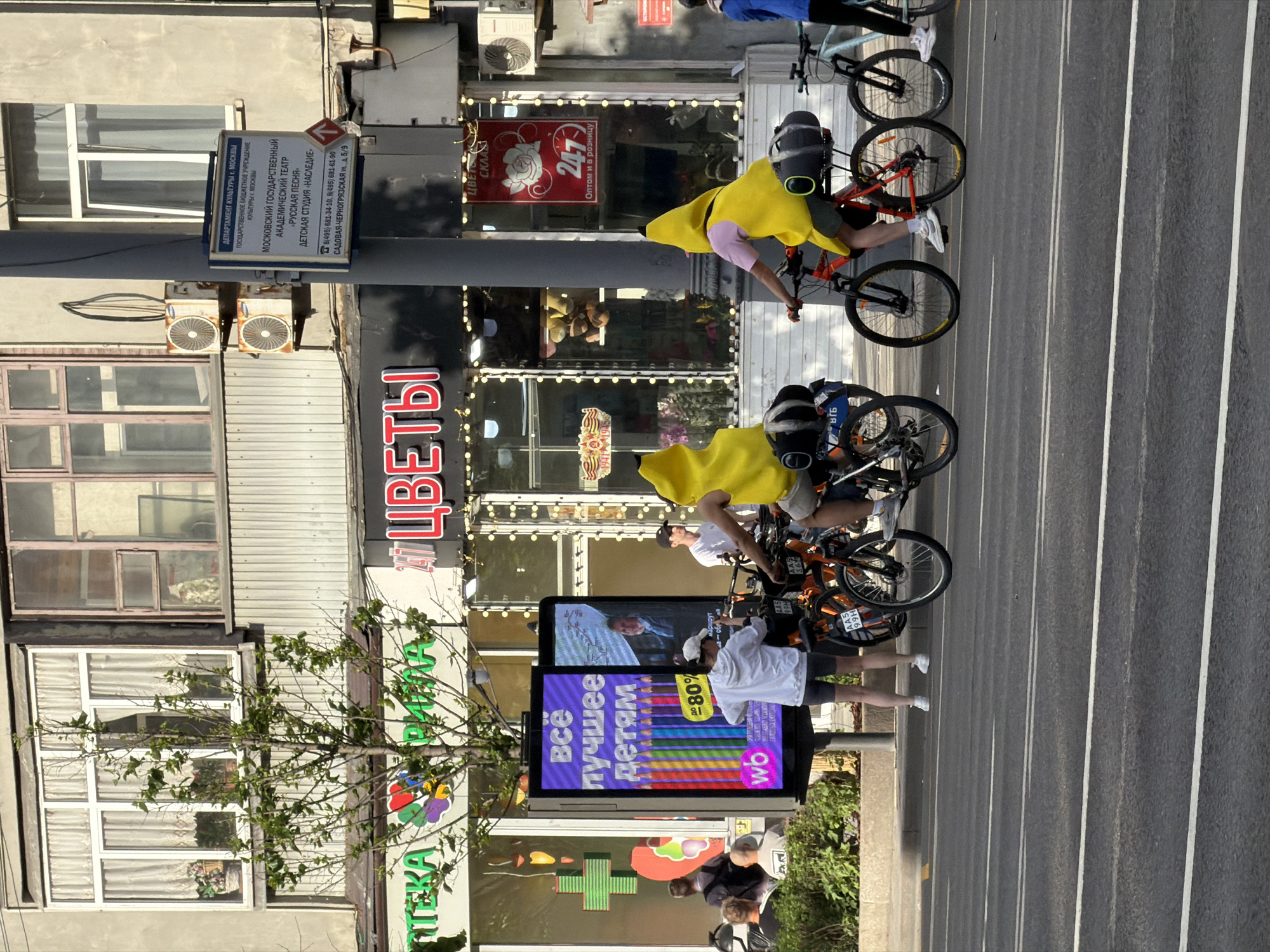 The image shows a street in Moscow, Russia. In the foreground are four cyclists riding on the road. Two of the cyclists are wearing banana costumes. The cyclists are riding past a sidewalk display promoting children's products at 80% off with the wildberries (wb) logo. Behind the display is a pharmacy (Аптека) with a sign, and a flower shop (Цветы) further back. A sign is visible on a lamppost that identifies the location as being near the Moscow State Academic Theater "Russian Song" Children's Studio "Heritage" at 5/9 Sadovaya Chernogryazskaya Street. Above the flower shop is a 24/7 sign (with a red rose) indicating that the shop is open 24 hours a day, 7 days a week, and offers both wholesale and retail services. The building in the background has multiple windows and air conditioning units. The street is mostly paved and painted with white lane markers.