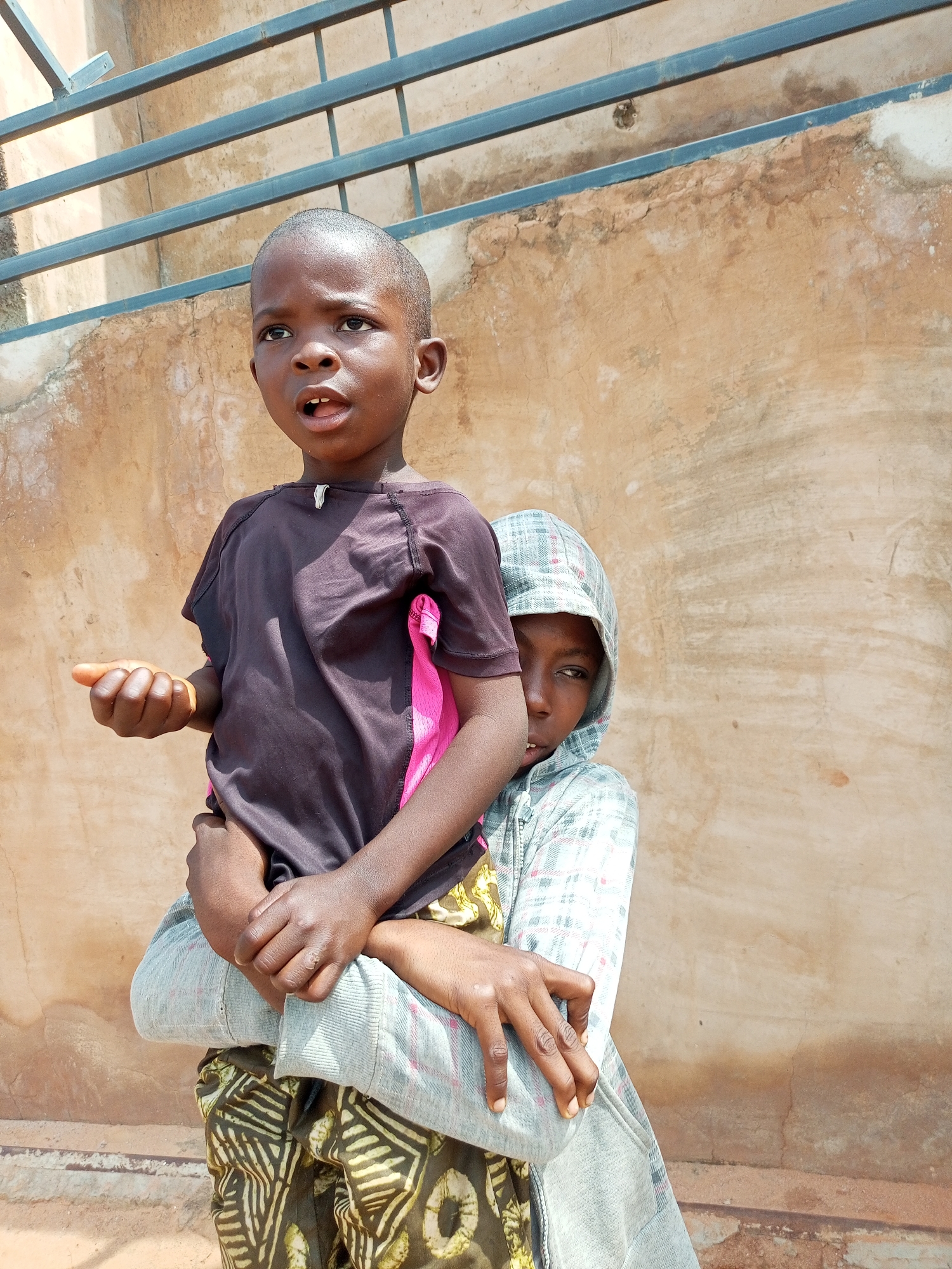 Two young children are standing in front of a textured, light brown wall in Tempure, Nigeria. The child in the foreground is facing left and looking upwards with their mouth slightly open. They are wearing a dark t-shirt with pink accents. Their left arm is bent and their hand is open. The second child is behind the first, embracing them from behind and is partially visible on the right side of the frame. This child is wearing a grey hooded sweatshirt and is looking to the left. Behind the children, a metal railing runs horizontally against the wall, and the ground is visible at the bottom of the image.