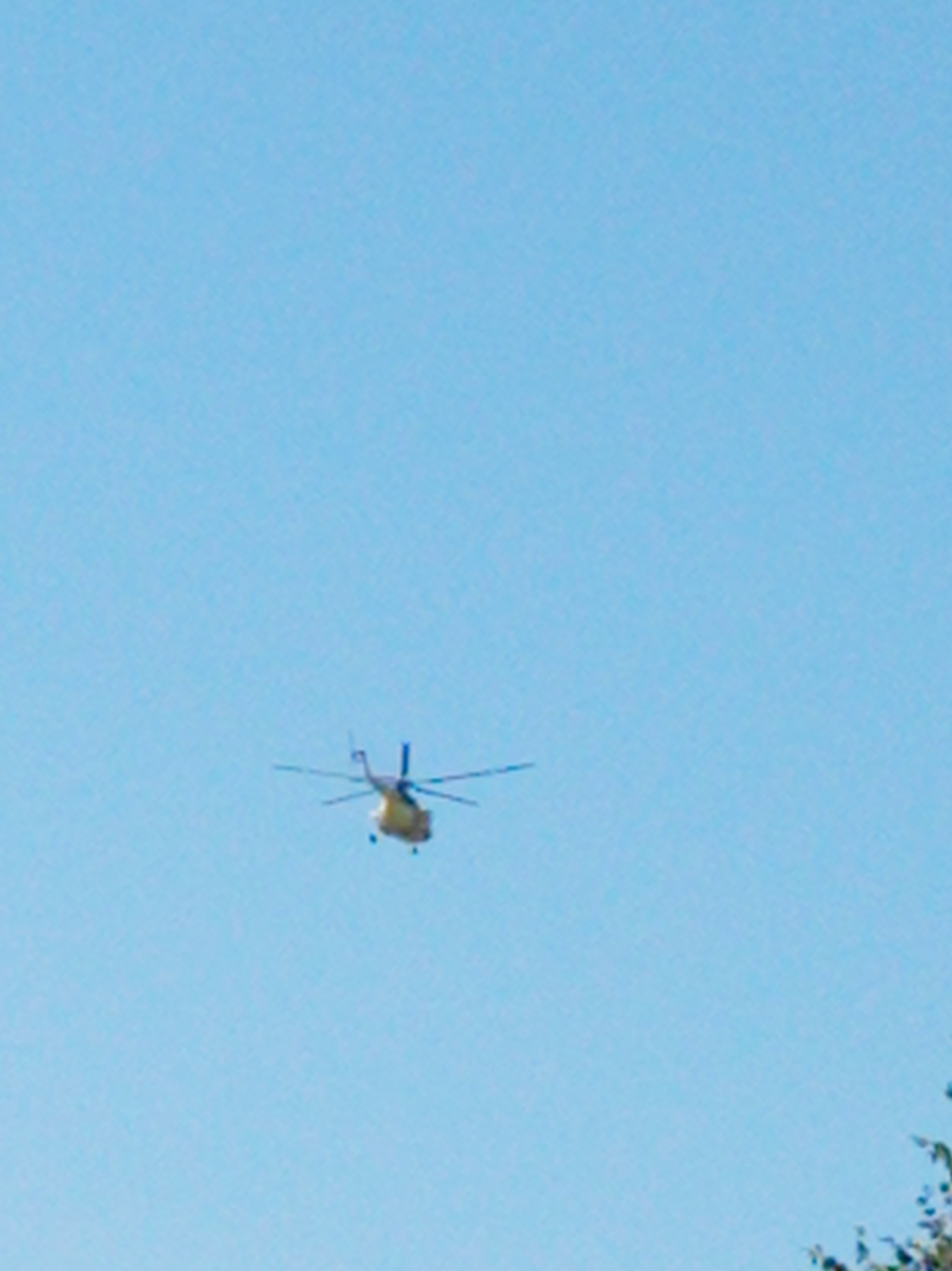 An aerial view shows a yellow helicopter flying in a clear blue sky. The helicopter is positioned in the center-left of the frame and is seen from below, with its rotors in motion. The background is an uninterrupted expanse of sky. There is no visible indication of the city of Artyom, Russia, within the image.