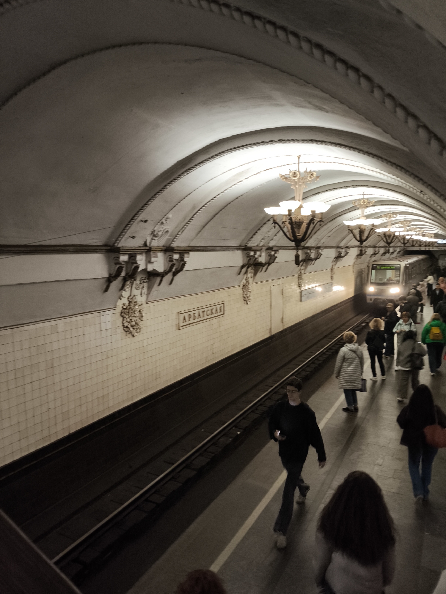 The image shows the platform of the Arbatskaya metro station in Moscow, Russia. A train is arriving. Several people are standing and walking on the platform. The station's architectural details, including arched ceilings, ornate chandeliers, wall decorations, and tiled walls, are visible.