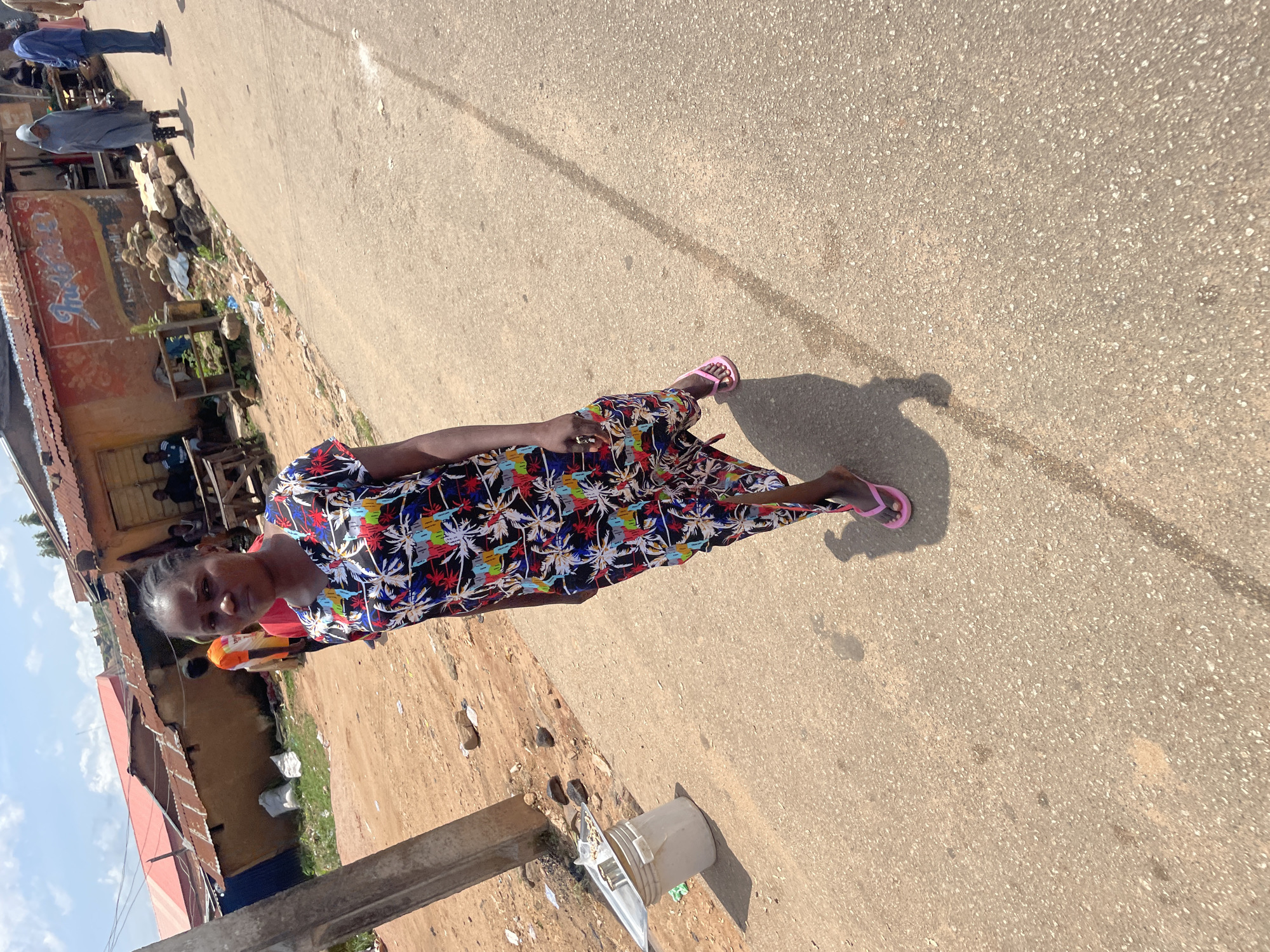 A young woman with braided hair, wearing a black dress with a tropical palm leaf print and pink flip-flops, walks on a paved road in Jos, Nigeria. The road is lined with informal buildings constructed with corrugated metal roofs and brick or concrete walls. To the left of the woman, there is a rough dirt area with scattered debris, including a white plastic bucket with a metal tray on top. In the background, several people are visible near the buildings, some seated on wooden benches. The sky is cloudy.