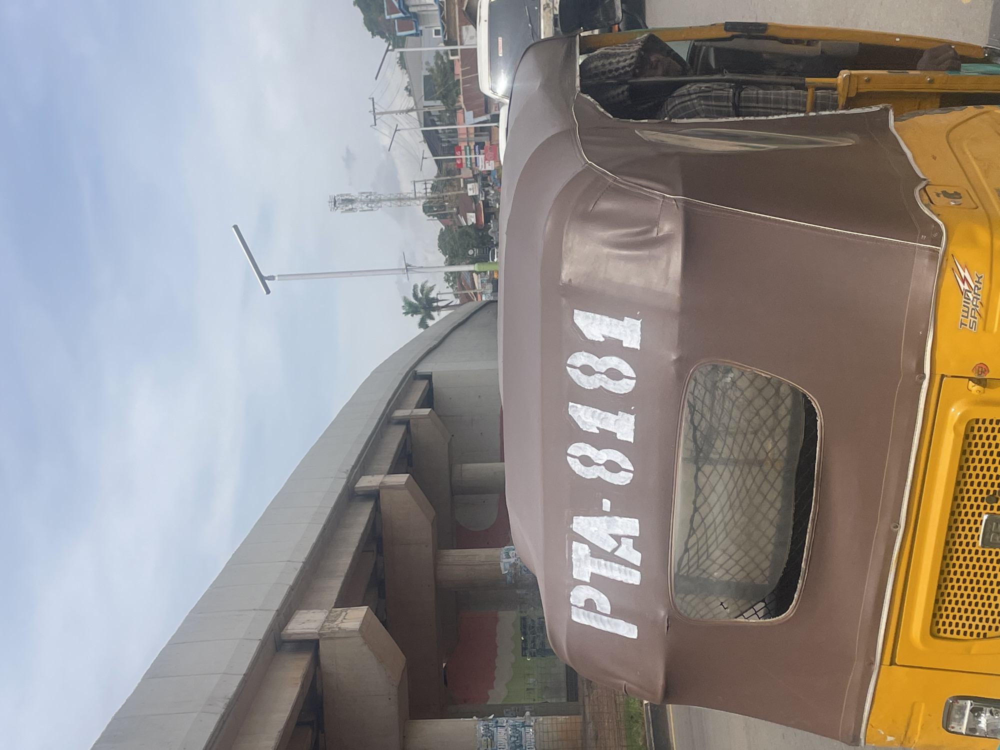 An outdoor, eye-level shot captures a portion of a yellow and brown auto-rickshaw in the foreground, identified by the white stenciled text "PTA-8181" on its brown canopy. A person wearing a checkered shirt and a beanie is visible inside the rickshaw. Behind the auto-rickshaw, a concrete overpass dominates the upper left of the frame, with its structural supports visible. To the right of the overpass, a street scene unfolds under a cloudy sky. In the mid-ground, a cell phone tower stands tall, flanked by streetlights and power lines. Buildings of various sizes and colors line the street, some adorned with signage. Traffic, including a white SUV, is present on the road. The overall setting suggests an urban environment, likely in Jos, Nigeria, given the presence of auto-rickshaws and the architectural style.