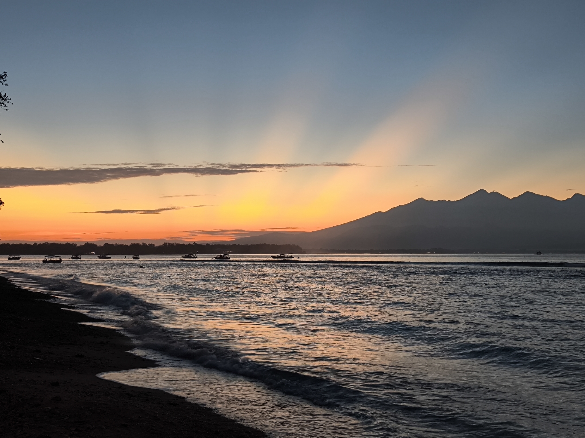 The image shows a sunset over a body of water, likely the ocean, in Unknown, Indonesia. The sky transitions from blue to orange, with distinct sun rays emanating from the horizon. A mountain range is visible in the distance, silhouetted against the sunset. Small boats are scattered on the water's surface. The foreground features a dark, sandy beach with small waves breaking on the shore. There is minimal vegetation visible in the scene.