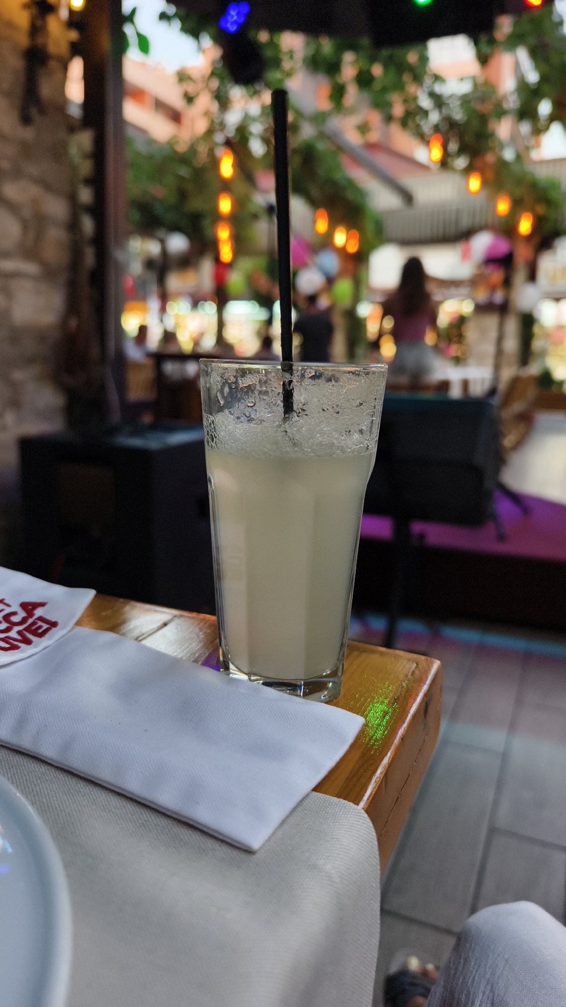 A tall, clear glass filled with a pale, frothy beverage and a black straw stands on a polished wooden table. The glass is slightly beaded with condensation, and the beverage appears to be a creamy cocktail or smoothie. In the foreground, a white folded napkin rests on the table to the left of the glass. The background is blurred, showing an outdoor setting with string lights, greenery, and patrons seated at tables, suggesting a lively atmosphere, possibly a cafe or restaurant in Kuşadası, Turkey. The overall lighting indicates it's either dusk or early evening.