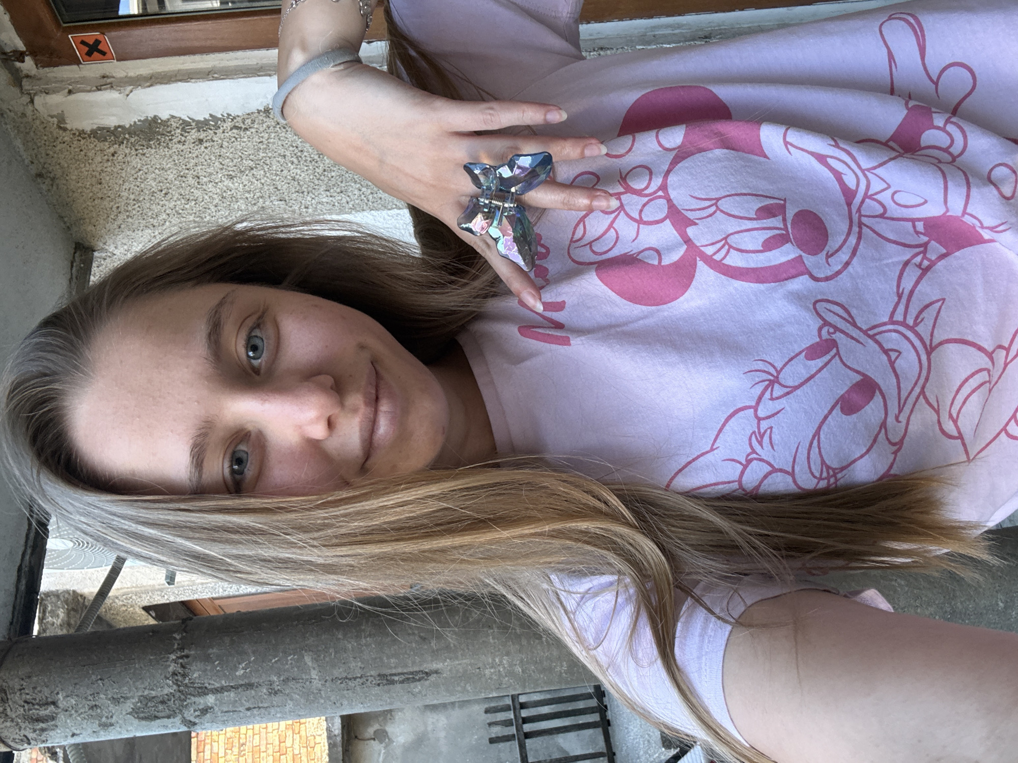 A young woman with long blonde hair and blue eyes is taking a selfie outdoors. She is wearing a pink t-shirt with Disney characters Minnie Mouse and Daisy Duck printed on it. She is also wearing a large, iridescent butterfly-shaped ring on her right hand and a grey scrunchie on her wrist. The background appears to be a courtyard or outdoor area with a concrete pillar, a brick wall, and part of a wooden door with a warning sign. The setting is consistent with an exterior area in Tsarevo, Bulgaria.