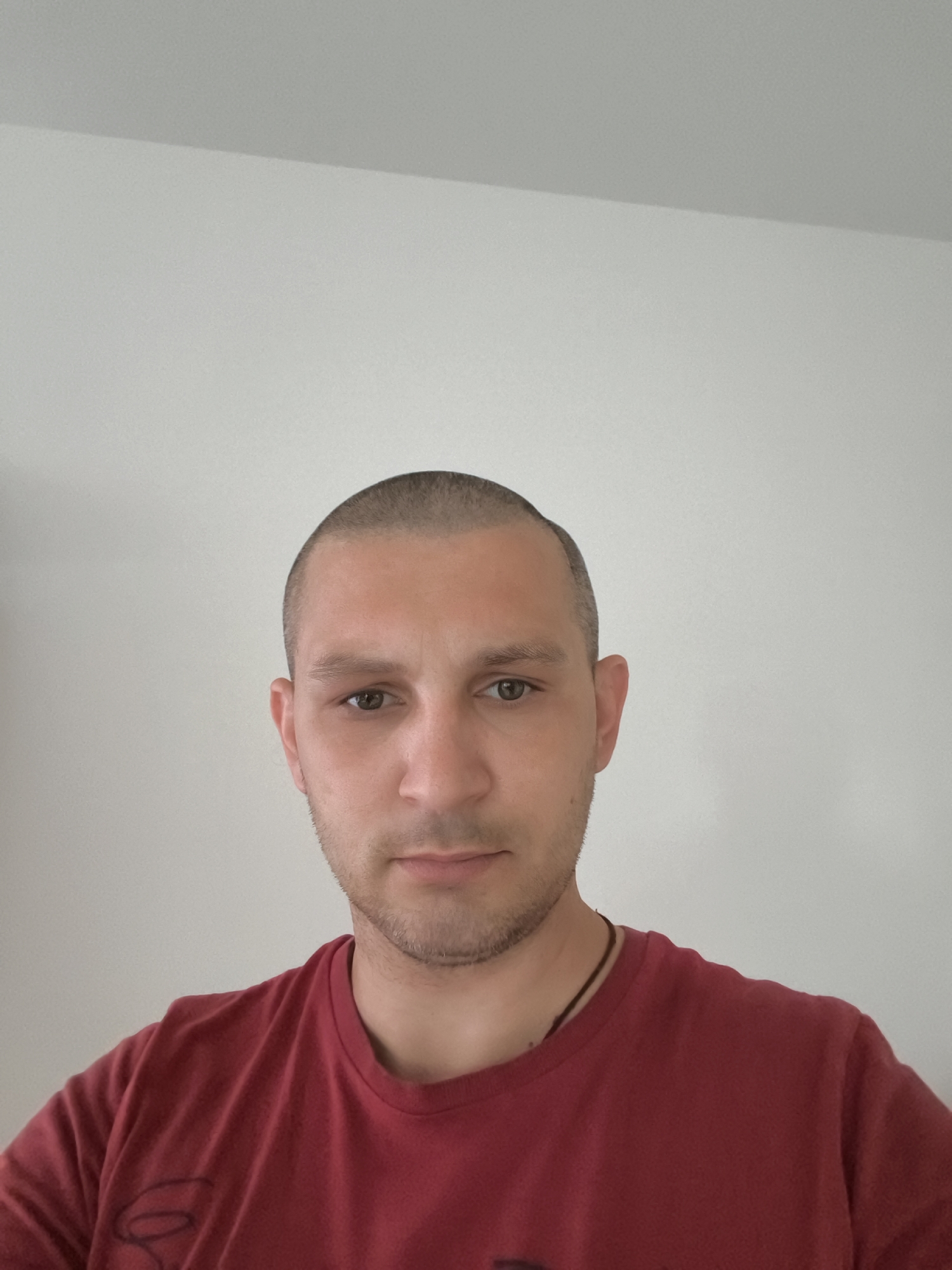 A man with a shaved head, light skin, and brown eyes is facing the camera. He is wearing a red t-shirt. The background is a plain white wall. The image was taken in Wolgast, Germany.