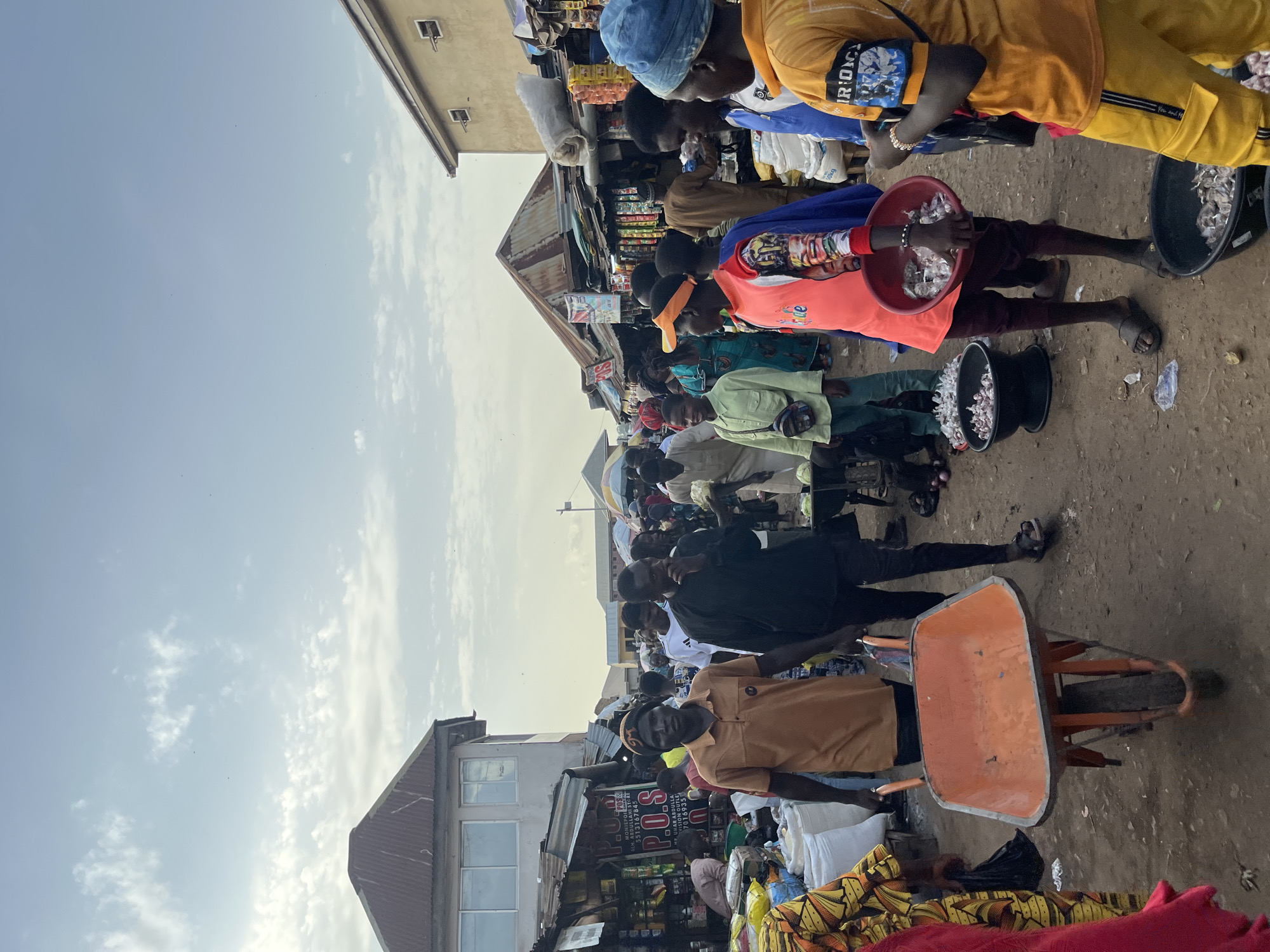 This is a photograph taken in Rafin Par, Nigeria. The scene depicts a bustling market with many people gathered. The subjects are predominantly young men and boys, dressed in casual attire, including t-shirts and trousers. Some are wearing hats or headscarves.
Objects visible include a wheelbarrow, several buckets (one red and one black) filled with what appears to be produce, possibly onions or garlic, and bags of goods. Stalls with various items for sale are set up, indicated by shelves stocked with packaged goods. The ground is dirt and some debris is scattered around.
The activity appears to be commerce, with people engaged in buying and selling. Some individuals are looking at the camera, while others are focused on their tasks or interacting with each other. There's a sense of a lively marketplace.
Notable details include the clear sky with some scattered clouds, suggesting it is daytime, likely late morning or afternoon. The buildings in the background appear to be made of concrete and corrugated metal, typical of market structures. The overall atmosphere seems busy and active.
Visible text includes "POS" prominently displayed on a sign, along with other inscriptions in what appears to be English and possibly another local language. There are also mentions of names like "ACH. ABDULLA" and "MR. UMAR ABDULLA," along with phone numbers. The text "50kg" and "IRONM" are also visible. The image is oriented vertically.