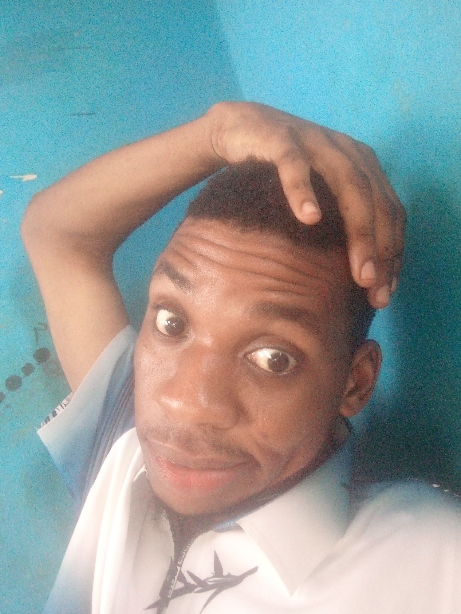 A medium close-up selfie of a young man with dark skin and short black hair. He is wearing a white collared shirt with a faint design on the chest, possibly a lightning bolt. His right hand is raised and resting on top of his head. He is smiling slightly with wide eyes. The background is a blue wall. The photograph was likely taken in Ikorodu, Nigeria, although there are no identifying elements in the picture to independently confirm this location.