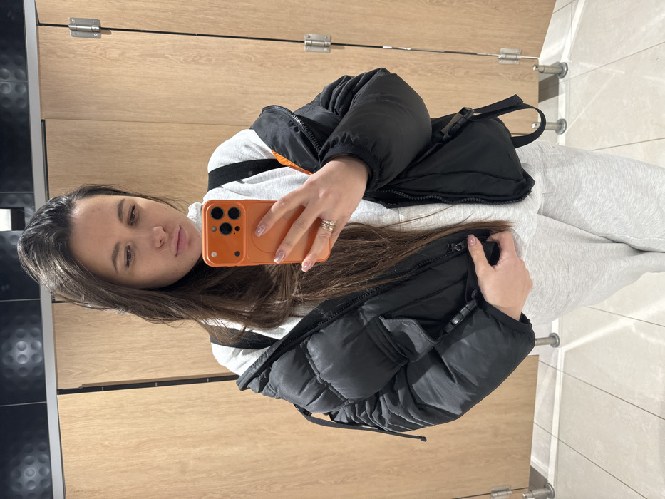 A woman takes a selfie in a restroom. She wears a gray tracksuit with a black jacket and a matching black bag. Her phone has an orange case. She is in front of light brown wood-paneled walls and a black tiled wall. A gray tiled floor is visible. The restroom includes metal fixtures and door hardware. She wears silver rings on her fingers.