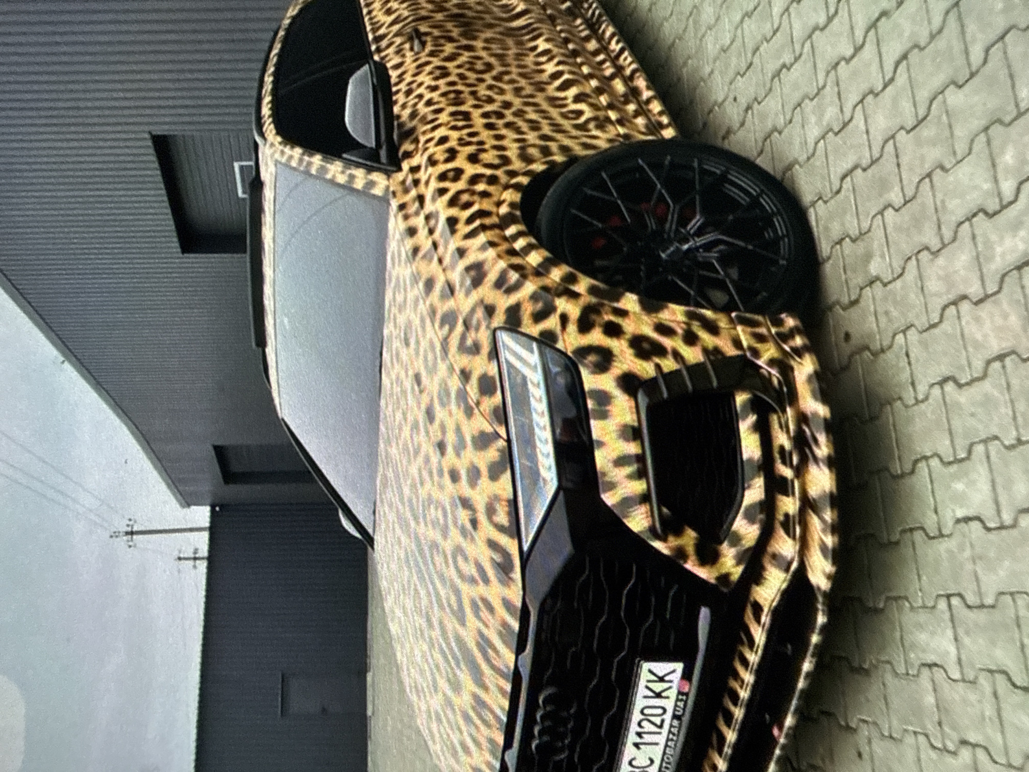 The image shows a car with a leopard print wrap, license plate "BC 1120 KK", parked on a paved surface. The location is possibly in an urban or suburban area. The surrounding buildings and power lines suggest a developed environment.