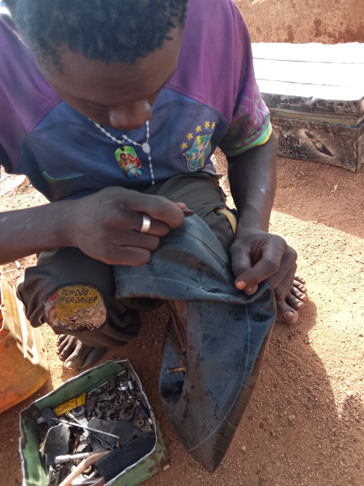 An individual with dark skin, wearing a short-sleeved shirt with purple and blue fabric, is repairing a tire tube. The person is crouched on reddish-brown dirt. On the left leg of the person's shorts is a sticker that reads "TOP DOG VIPER DRIVER." A container filled with metal objects sits to the person's left. The scene is situated in Jos, Nigeria.