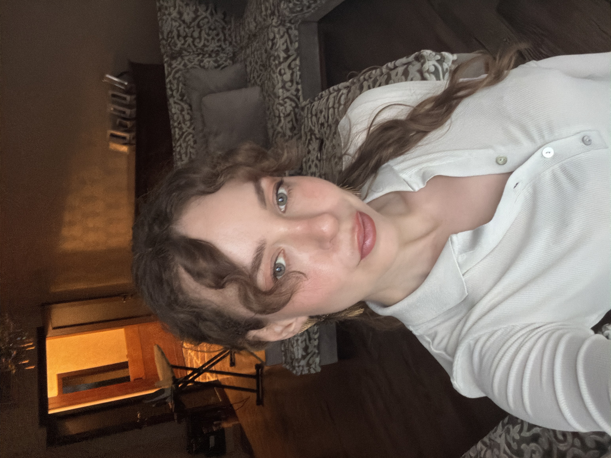 A young woman is captured in a close-up selfie, looking directly at the camera with a calm, pleasant expression and a slight smile. Her curly, light brown hair frames her face, with some strands falling across her forehead. She has prominent blue-green eyes and her lips are painted a soft pink. She is wearing a white, collared, long-sleeved top with buttons down the front.
The setting appears to be indoors, likely a living room or bedroom, characterized by warm, dim lighting. Behind her, a patterned sofa or bed with decorative cushions is visible, featuring a dark brown and beige intricate design. To the left side of the frame, a dark wooden shelf or cabinet is present, from which an orange glow emanates, suggesting an internal light source or reflection, contributing to the cozy atmosphere. Below this, a structure with black metal bars, possibly part of furniture like a bed frame or side table, is discernible. The overall ambiance is relaxed and intimate, indicative of an evening or nighttime setting. No specific visual cues confirm the location as Moscow, Russia, beyond the provided context. There is no visible text in the image.