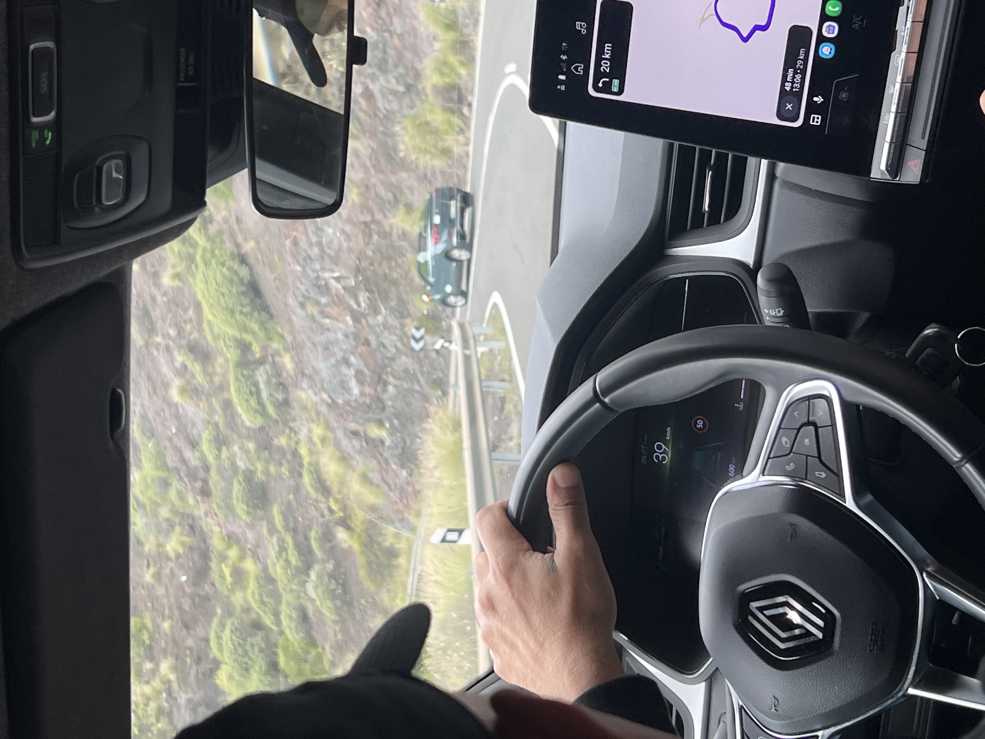 The image, taken from the driver's seat, captures a scene of driving along a winding road in Mogán, Spain. The driver's hands are on the steering wheel of a Renault, indicated by the logo. The dashboard screen shows a speed of 39 km/h. A navigation app is displayed on a tablet mounted on the dashboard, indicating the destination is 20 km away and the estimated time of arrival is 48 minutes later at 13:06. Outside the window, the landscape reveals a mountainside covered with greenery. A vehicle is visible on the road ahead.