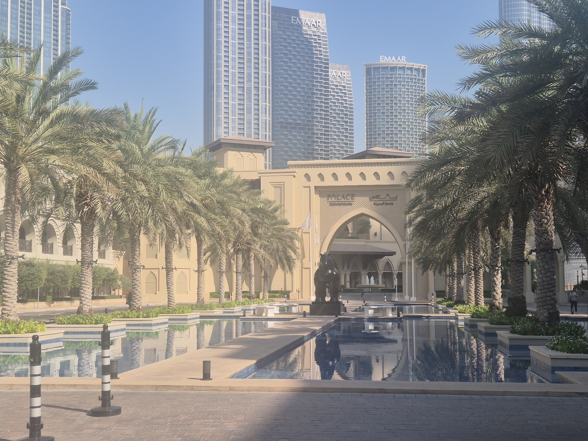 The image shows an outdoor scene in a modern urban environment. In the foreground, there is a paved walkway with several black and white striped bollards. Beyond the walkway, a shallow pool with a tiled bottom is visible, reflecting the surrounding structures and palm trees. Several planters with greenery are positioned along the edge of the pool.
In the midground, a row of mature palm trees stands tall, their fronds casting shadows. Behind the palm trees, there is a tan-colored building with arched doorways and decorative patterns. A large, dark sculpture of an animal, possibly a horse, is placed on a pedestal in front of one of the arches. The building appears to be an entrance to a larger complex.
In the background, several towering modern skyscrapers dominate the skyline. One of these buildings prominently displays the "EMAAR" logo. The sky is clear and blue, suggesting a sunny day. The overall impression is one of a well-maintained, upscale public space, likely in a city known for its contemporary architecture. The architectural style of the tan building suggests a blend of traditional Islamic design elements with modern construction. The presence of multiple palm trees and water features indicates a warm climate. The image is taken from a slightly low angle, looking up towards the skyscrapers. There is one person visible walking in the distance on the right side of the image.