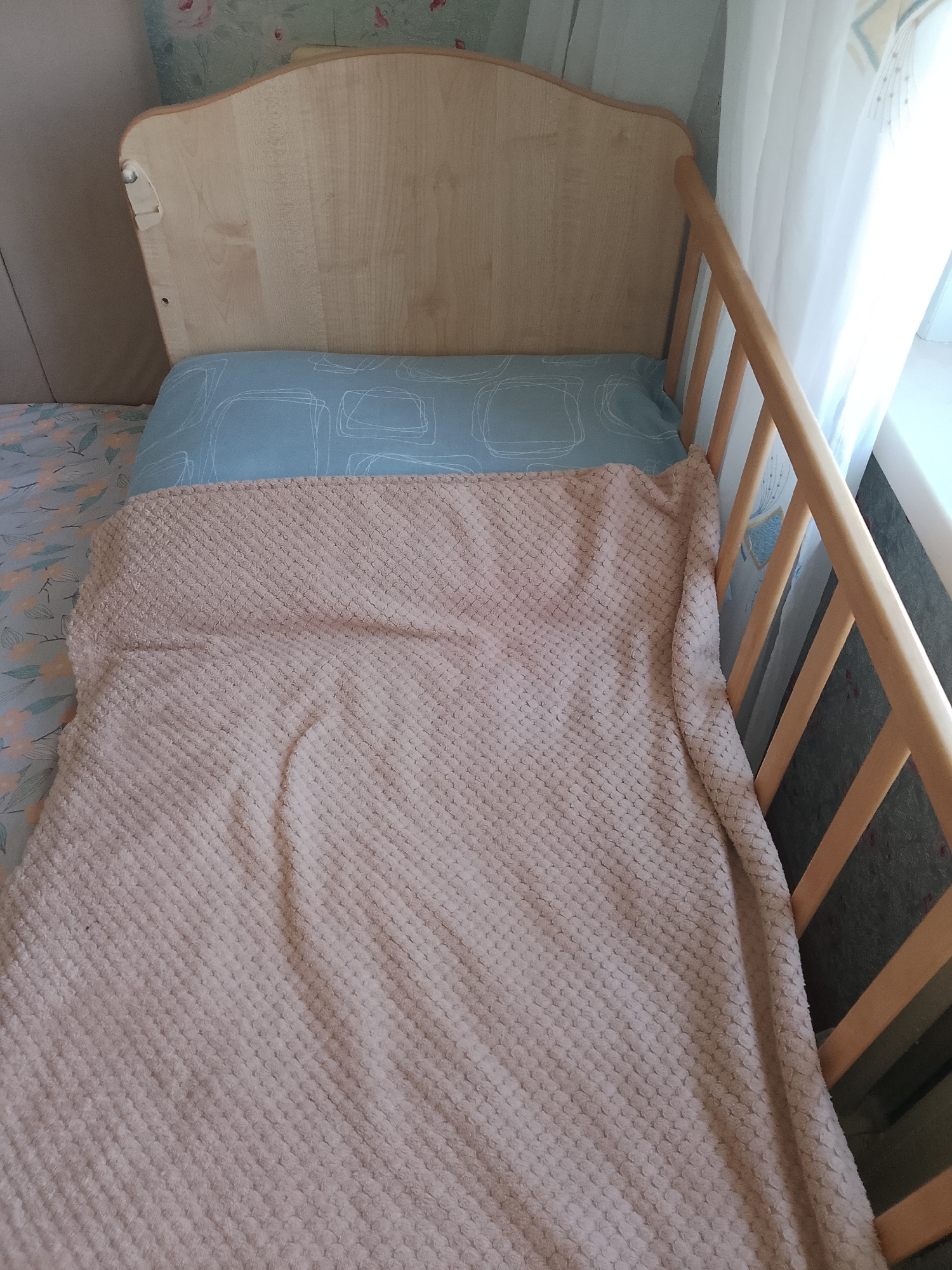The image shows a close-up view of a crib with a wooden frame. A light blue pillow with a white line pattern is visible at the head of the crib, covered by a beige textured blanket. The crib is positioned next to a window with sheer white curtains, and a wall with floral wallpaper is in the background. The lighting suggests daytime. The image does not provide information about the specific location within the city of Abay, Kazakhstan.