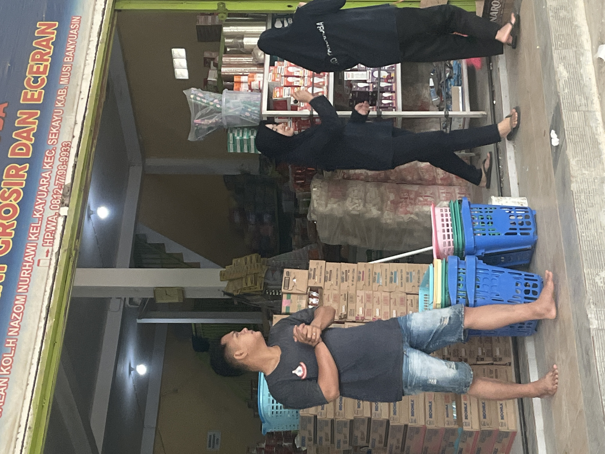 The image depicts the exterior of a storefront in Sekayu, Indonesia, indicated by text on the signage which reads "JL. KOL.H. NAZOM NURHAWI KEL. KAYUARA KEC. SEKAYU KAB. MUSI BANYUASIN HP. WA. 0812-7798-9933". The storefront appears to be a wholesale and retail establishment, with various goods displayed.
In the foreground, a man is standing to the left, facing right. He is wearing a dark t-shirt and denim shorts, and is barefoot. His hands are clasped in front of him.
To the right, two women in dark headscarves and long dark clothing are interacting with the store's inventory. One woman is reaching for an item on a shelf, while the other stands with her back to the camera. The shelves are stocked with various products, including what appear to be light bulbs and other packaged goods.
Behind the man, stacks of cardboard boxes are visible, some with labels. In front of the shelves, there are several blue plastic laundry baskets, some with colorful plastic lids.
The environment suggests a commercial area, with the building having an open-front design allowing visibility into the store's interior. Lighting fixtures are visible on the ceiling inside the store. The overall impression is one of an active retail space in a town.
