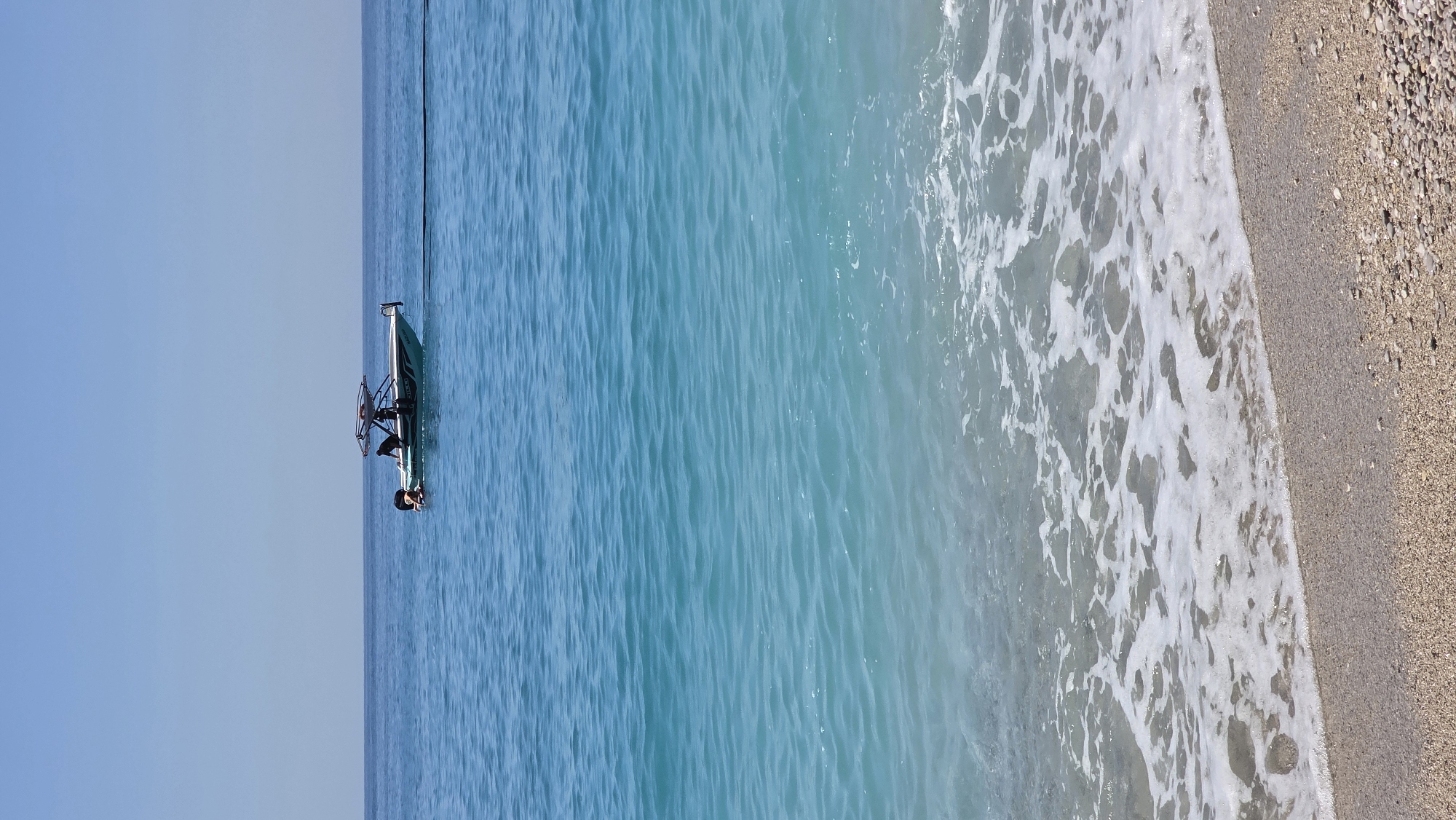 The image displays a serene coastal scene in Fethiye, Turkey, under a clear, bright sky. The perspective is from the shore, looking out over the sea towards the horizon.
The upper-left portion of the image is dominated by a vast, cloudless light blue sky. Below it, stretching across the majority of the frame, is the vibrant turquoise sea. The water appears calm and clear in the distance, gradually becoming lighter and showing gentle white foamy waves as it approaches the shore.
In the middle-left section of the calm sea, a small, light-colored watercraft, possibly a pedalo or a small catamaran, floats stationary. It features a simple framework structure. Two individuals are present with the vessel: one person is seated or leaning on the craft, and another is partially submerged in the water beside it, with their head and shoulders visible. Both individuals have dark hair and appear to be engaged in leisure activities.
On the far right, a narrow strip of pebbly beach runs vertically. It consists of light brown and beige stones and pebbles, where the foamy edge of the breaking waves gently meets the land.
The scene conveys a peaceful, leisurely atmosphere, characteristic of a sunny day by the Mediterranean. There is no visible text in the image.