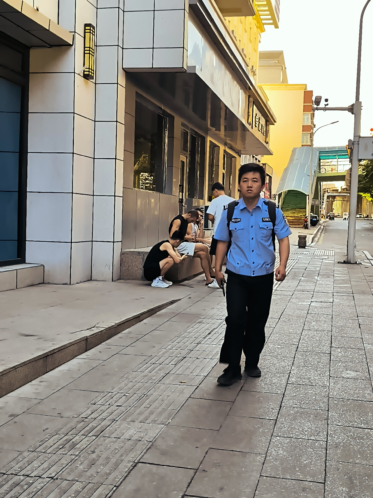 A young East Asian man in a light blue uniform shirt and black pants walks on a city sidewalk. The man is looking forward, and has short black hair. He has a black backpack on. A badge is visible on his shirt. The sidewalk is paved with light gray square tiles. To the man's left, there is a building with a white tiled facade. Several other people are seated on a stone bench in front of the building, looking at their phones. To the man's right, the sidewalk continues, and there is a street with a pedestrian overpass in the distance. The sun is shining, casting long shadows. This scene is consistent with urban environments in Ürümqi, China.