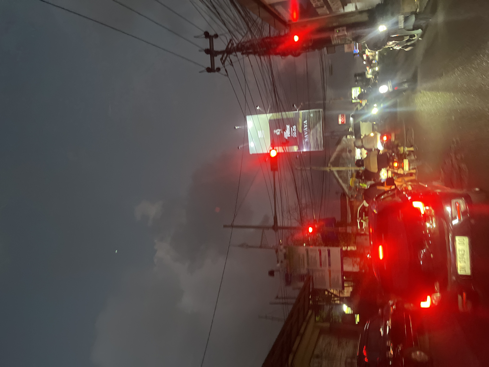 The image depicts a street scene in North Kuta, Indonesia, at dusk or night, characterized by heavy traffic and a gloomy sky. The foreground is dominated by the red taillights of a black car, with its license plate visible as "DM 2843 DU". Several other vehicles and motorcycles are present in the traffic jam, many with their headlights illuminated, creating streaks of light.
In the background, a large billboard advertising "SAVAYA" is illuminated. Traffic lights are visible, with at least one showing red. A utility pole with numerous wires runs along the right side of the street, typical of urban infrastructure. The sky is overcast with dark clouds, and there appears to be a faint green light in the sky in the upper left portion of the image. The overall impression is one of a busy urban environment during adverse weather conditions or low light.