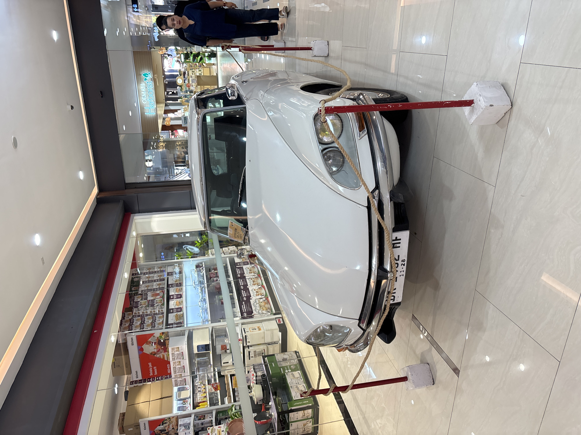 The image captures a vintage white car on display inside a shopping mall in Kuta, Indonesia. A red rope barrier held up by red poles and white cuboid bases surrounds the car. A sign is visible on the car's windshield, and its license plate is partially visible.
The backdrop reveals the interior of a department store with rows of shelves stocked with various kitchenware items and boxes. A glass partition separates the car display from the store's interior. Further back, a shop sign reading "OEMAH Herborist" is discernible. In the distance, two individuals are visible, suggesting customers strolling through the mall. The mall floor is tiled in a light color, and the lighting is bright and even.