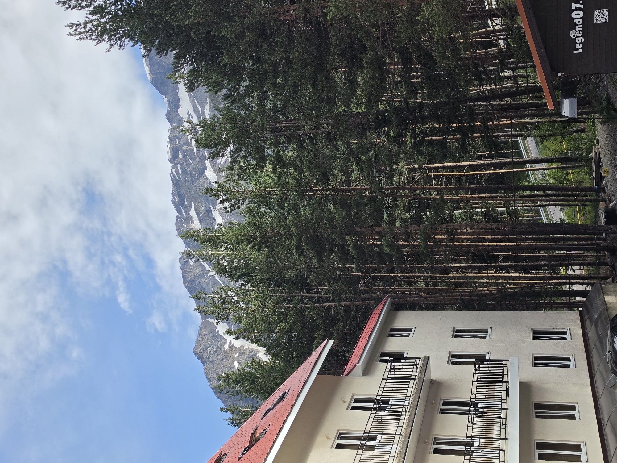 The image depicts a view of a building and surrounding natural landscape in Terskol, Russia. In the foreground on the left, a multi-story building with a red-tiled roof and cream-colored walls is visible. It features balconies with black railings and numerous windows. Behind the building, a dense forest of tall pine trees extends towards the background. The trees are a dominant feature, with their green foliage creating a textured wall. Further in the distance, a mountain range with snow-capped peaks rises against a partly cloudy blue sky. The overall scene suggests a location in a mountainous, forested area, likely a resort or residential area given the presence of the building. The lighting indicates it is daytime, with sunlight filtering through the trees.