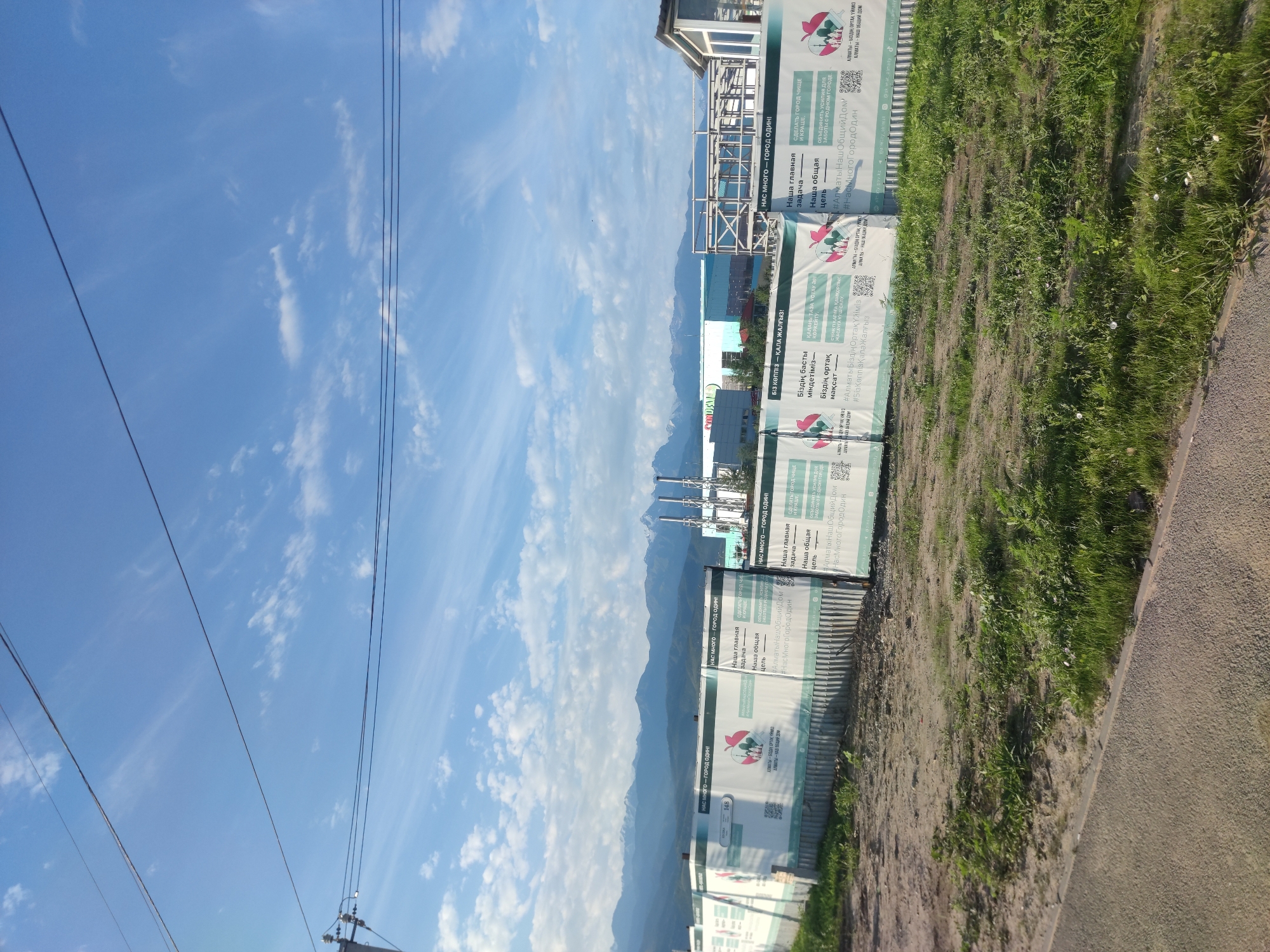 This image depicts a construction site in Almaty, Kazakhstan, with mountains in the background. A white banner with text and images is prominently displayed, likely related to a city improvement or development project. The banner has sections in both Kazakh and Russian, indicating a bilingual context. The text mentions "Наша главная задача" (Our main task) and "Наша общая цель" (Our common goal), along with hashtags such as "#АлматыНашОбщийДом" (#AlmatyOurCommonHome) and "#НасМногоГородОдин" (#WeAreManyCityOne). The construction site appears to be fenced off with metal barriers and a dirt ground area with sparse vegetation. In the far background, industrial structures with tall chimneys are visible, along with some buildings that appear to be commercial or residential. The sky is blue with scattered white clouds, and power lines are visible in the upper part of the frame.