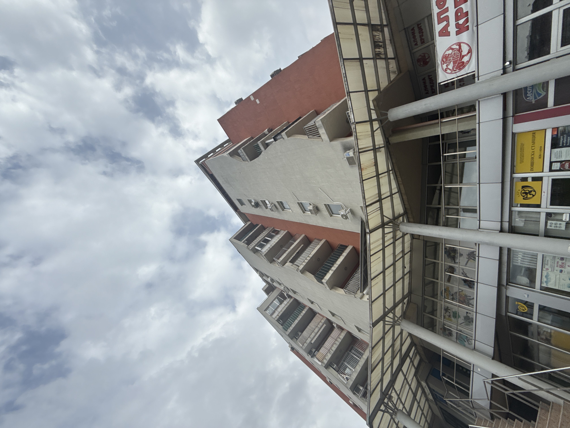 A multi-story building, likely a mixed-use commercial and residential structure, stands tall against a cloudy sky. The image is taken from a low-angle perspective, looking upwards. The building features several balconies with railings on its residential sections, painted in a light beige color, contrasted with a reddish-brown section higher up. Numerous air conditioning units are visible mounted on the exterior walls.
The lower section of the building appears to be commercial, with large glass windows and an awning structure extending over what might be a pedestrian area or entrance. Grey pillars support this overhead structure. A partial view of a stairwell with metal railings is also visible at the bottom right.
The weather is overcast, with a sky full of grey and white clouds, indicating daytime. No people or specific activities are discernible in the static scene.
Several Cyrillic texts are visible on banners and signs on the commercial facade. Prominently displayed are banners repeating "АЛФИА КРЕДИТ" (Alfia Kredit), suggesting a credit or financial service. Another sign reads "ПОЩЕНСКА СТАНЦИЯ 1836 софия" (Postal Station 1836 Sofia), clearly indicating a post office and confirming the location as Sofia. Other smaller texts include "Дест", "point", "1979", "1-07", "80", and text partially visible that might relate to "на оборота в България" (turnover in Bulgaria).