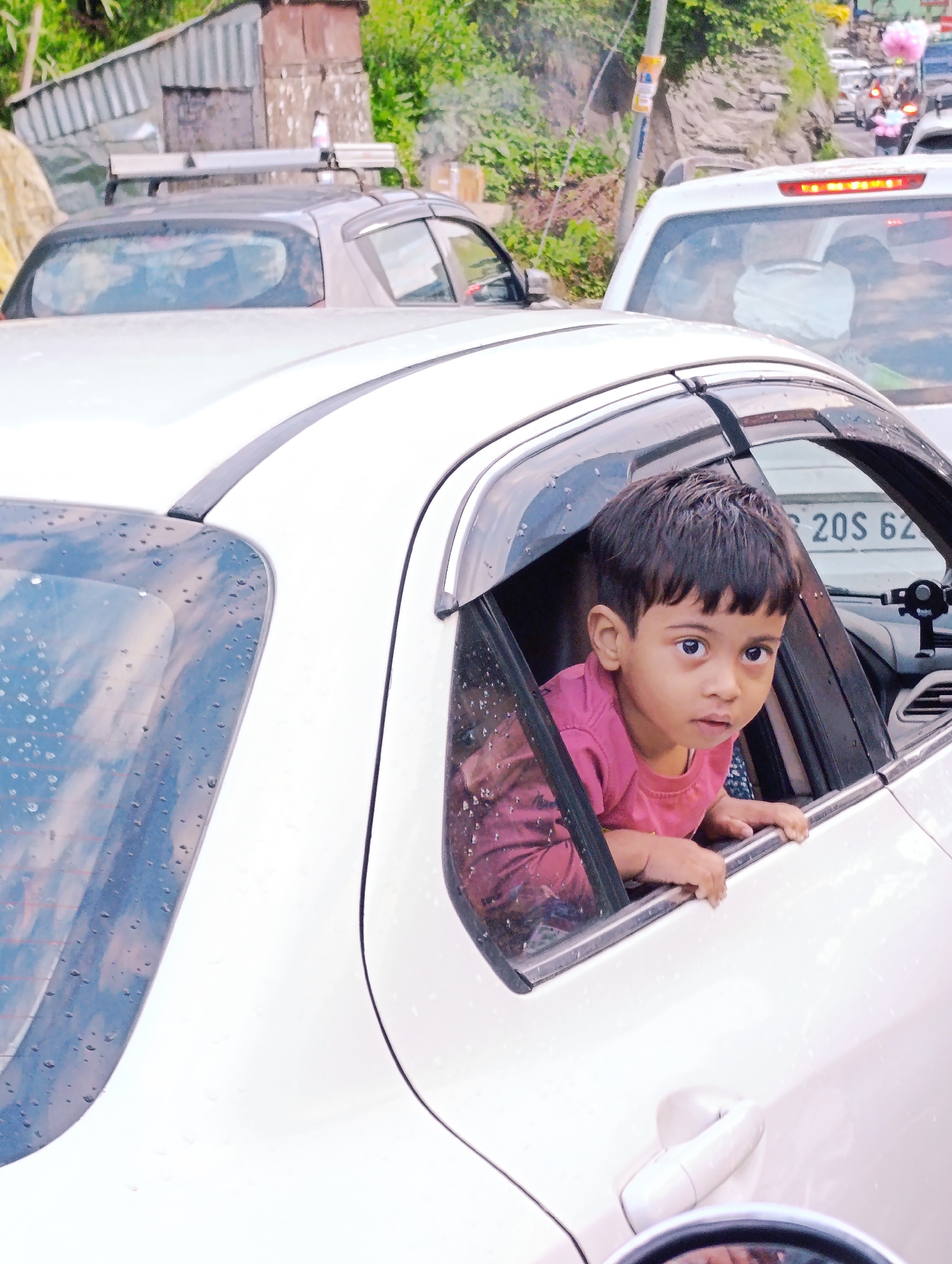 A young boy peers out of the window of a white car during a traffic jam in Kulang, India. The car's windows are speckled with water droplets, suggesting it may be raining or has recently rained. The boy is wearing a pink shirt and is holding onto the edge of the car window. In the background, other vehicles and a roadside structure are visible. The image captures a moment of stillness amidst the movement of traffic.