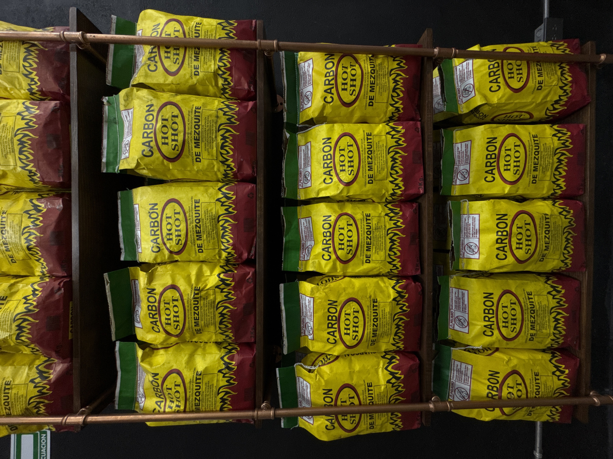The image shows a display of "Carbon Hot Shot De Mezquite" charcoal briquettes in a retail setting. The yellow and red bags are stacked on a dark wooden shelving unit with metal railings. The environment appears to be a store, possibly in Puebla, Mexico, given the Spanish on the packaging. There are no people visible. The bags are arranged in neat rows. The bags feature a prominent "Carbon Hot Shot" logo and branding, with information about the product's net weight and manufacturer.