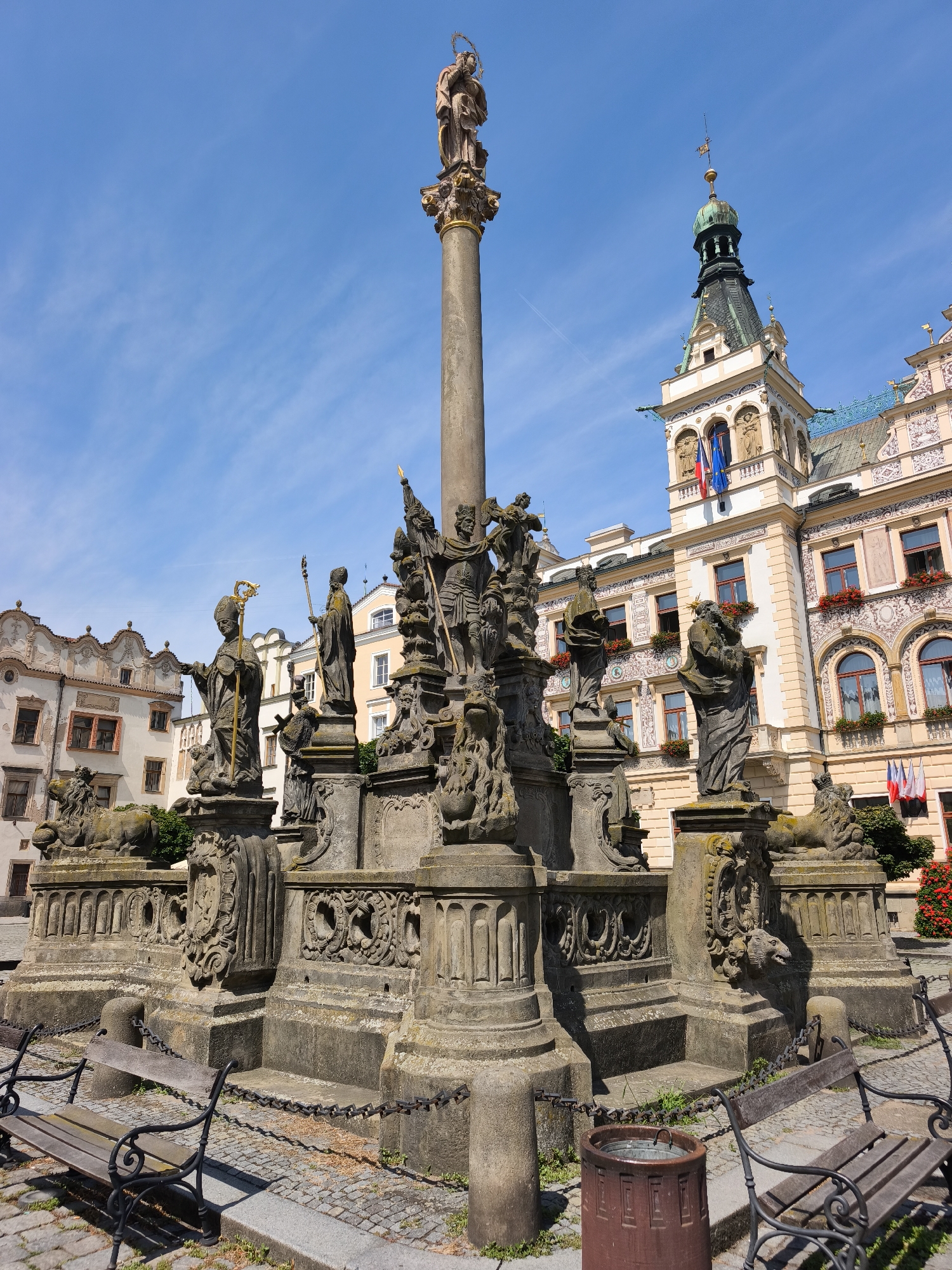 This media file captures a sunny outdoor scene featuring a Baroque plague column situated in a public square. The column is adorned with numerous statues of saints and mythological figures, and is made of weathered stone. Atop the column stands a statue of the Virgin Mary with the Christ Child, crowned with a golden halo. At the base of the column are stone lions, and the entire structure is surrounded by decorative stone carvings. Two empty park benches are visible in the foreground, suggesting a place for rest.
In the background, a grand, ornate building with a green, multi-tiered spire dominates the skyline. The building displays architectural details such as arched windows, decorative facades, and flags, including the Czech flag and a blue and yellow flag, likely representing the European Union. Red flowers are visible in window boxes, adding a splash of color to the scene.
The sky is a clear, bright blue with a few wispy white clouds, indicating pleasant weather. The lighting suggests it is daytime, likely mid-morning or early afternoon. The scene is static, with no active event taking place, giving a sense of peacefulness and historical grandeur. The location is identified as Pardubice, Czechia. There is no visible text in the media file.