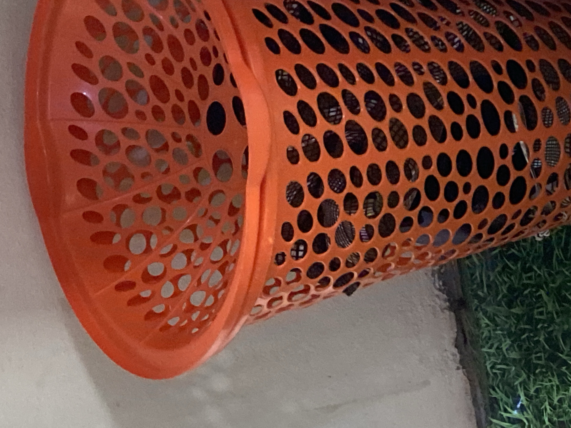 In Jauro Yinu, Nigeria, a close-up, eye-level shot captures a bright orange plastic laundry basket positioned against a textured, off-white wall. The basket is filled with what appears to be dark, patterned fabric. The lower portion of the basket extends to the right, obscuring the rest of the scene. The ground beneath the basket is covered with artificial green turf. The lighting is moderate, with some shadows visible on the wall. There are no people or animals in the frame.